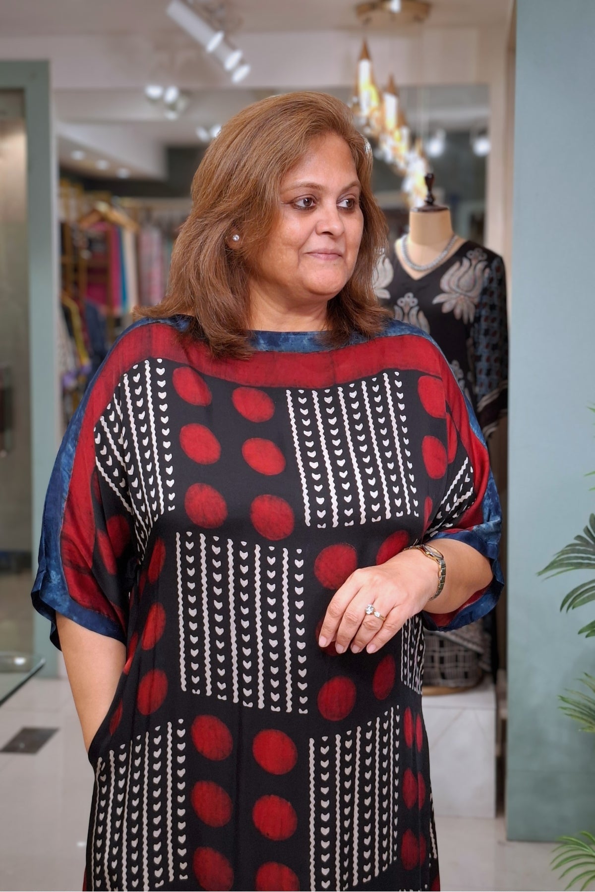 A woman wearing a loose-fitting black tunic dress with red circular patterns and white vertical geometric stripes, standing in a boutique.
