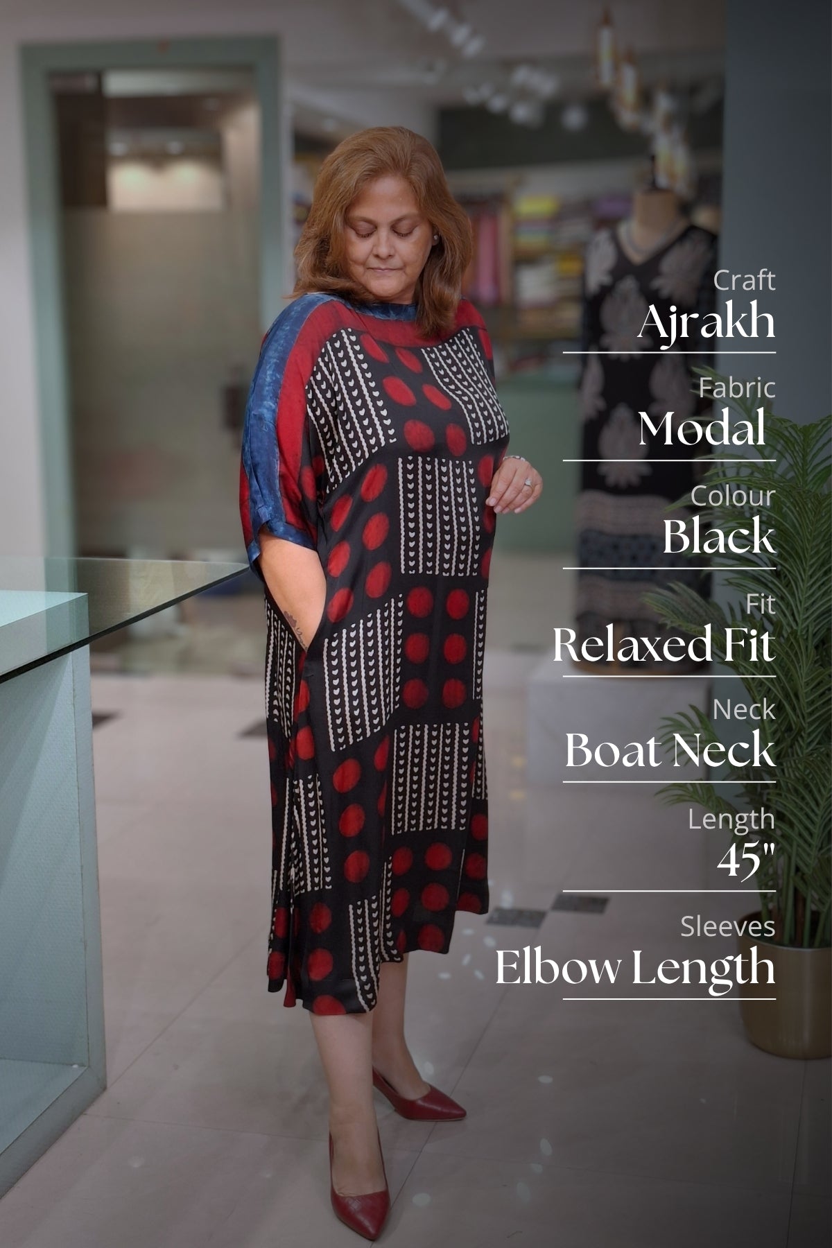 A woman modeling a black, red, and blue Ajrakh fabric dress in a boutique setting, with text overlays detailing the dress specifications on the right side.