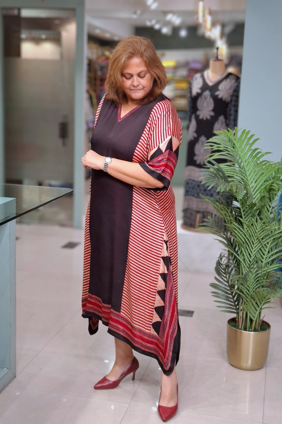 A woman wearing a long, loose-fitting kaftan dress with a color-blocked design of solid black, red, and beige stripes, standing in a boutique next to a potted plant.