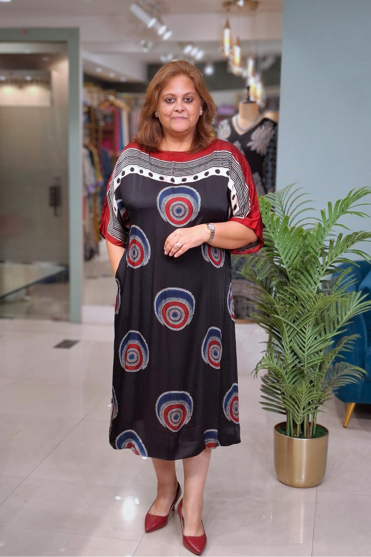 A woman modeling a black midi dress with red, blue, and white circular patterns, standing in a boutique next to a potted plant.