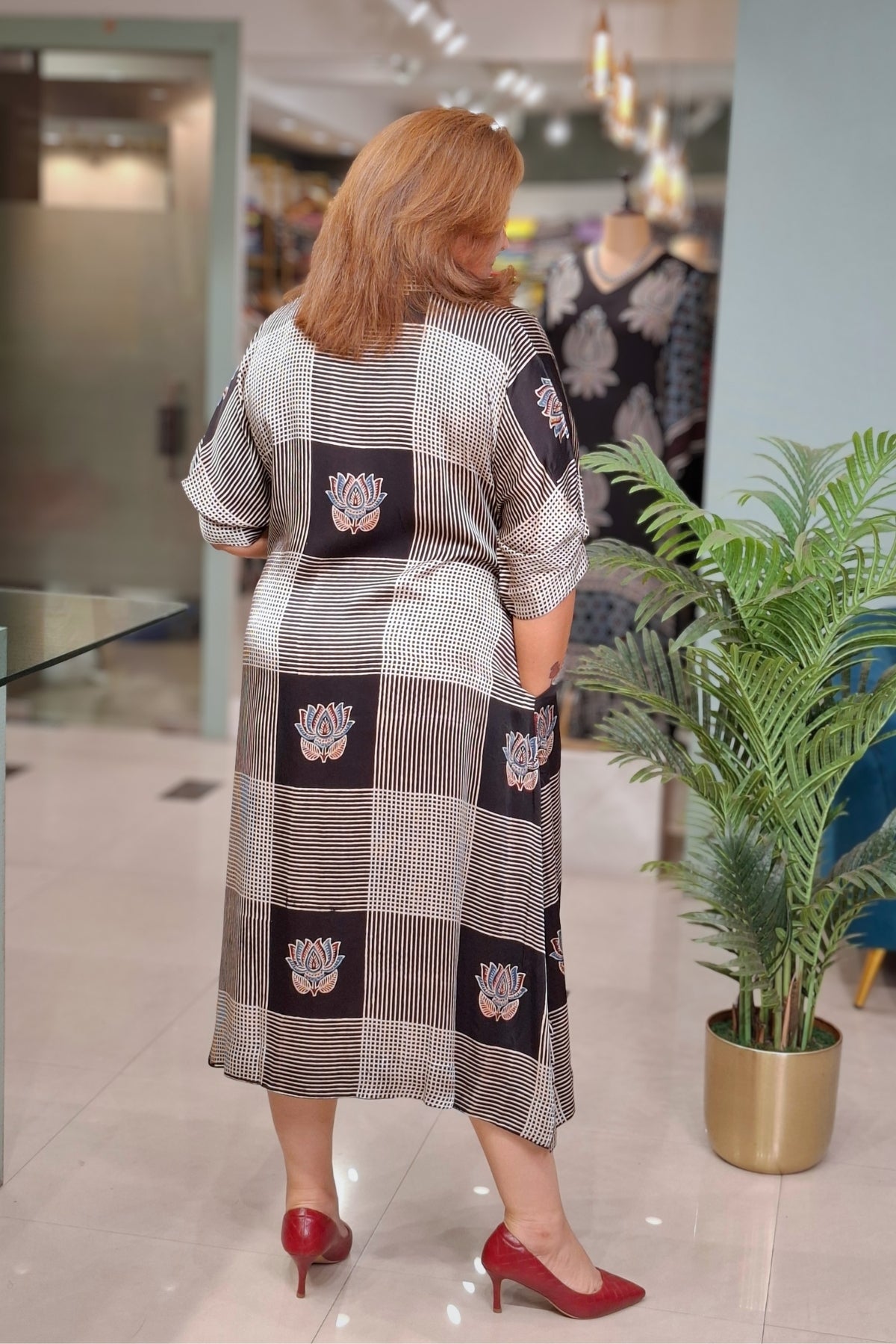 A woman viewed from behind wearing a long-sleeved, knee-length checkered dress with lotus motifs in a boutique setting, paired with red heels.