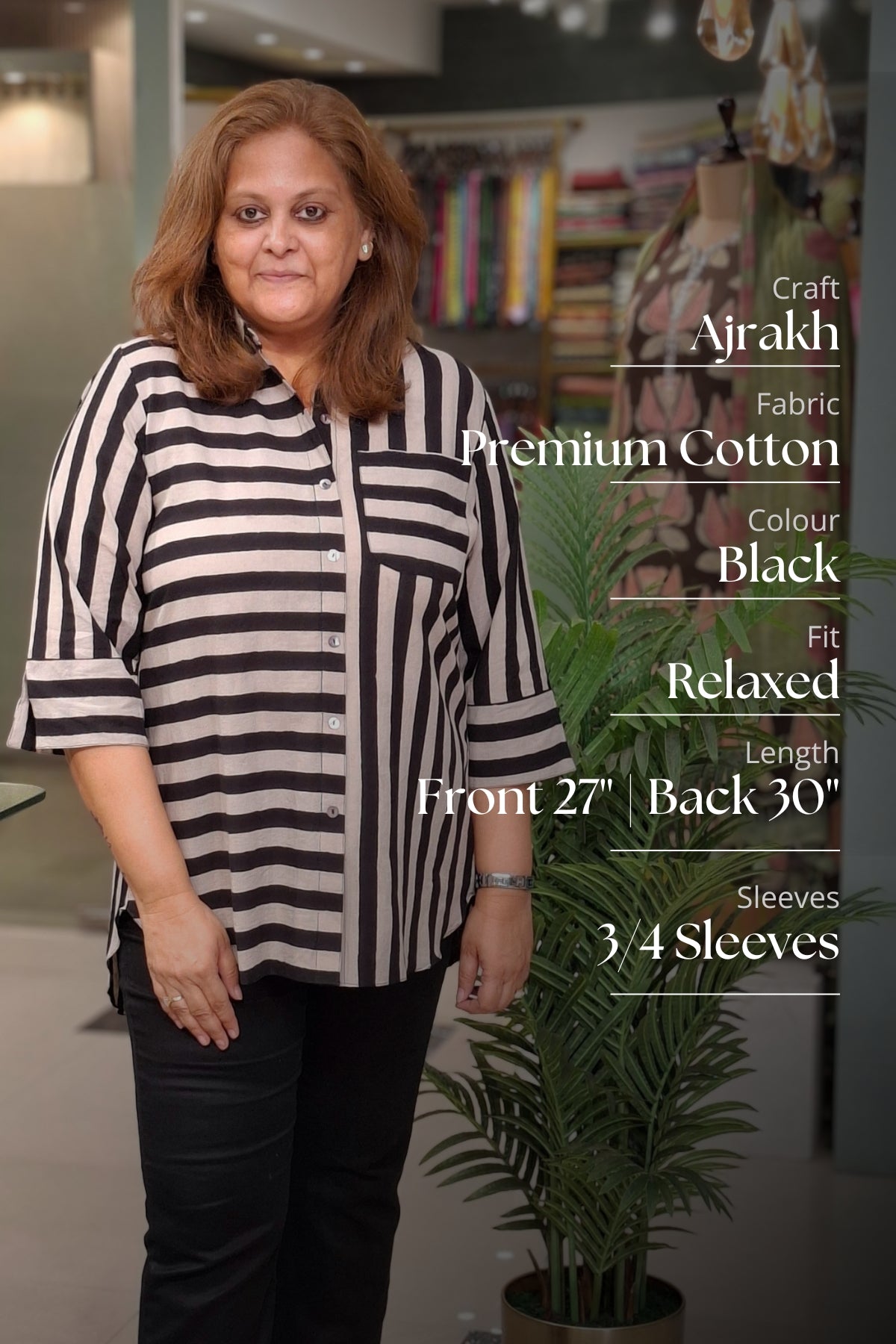 A woman modeling a black and white striped relaxed-fit cotton shirt in an indoor boutique setting, with product text overlays on the right detailing fabric and fit specifications.