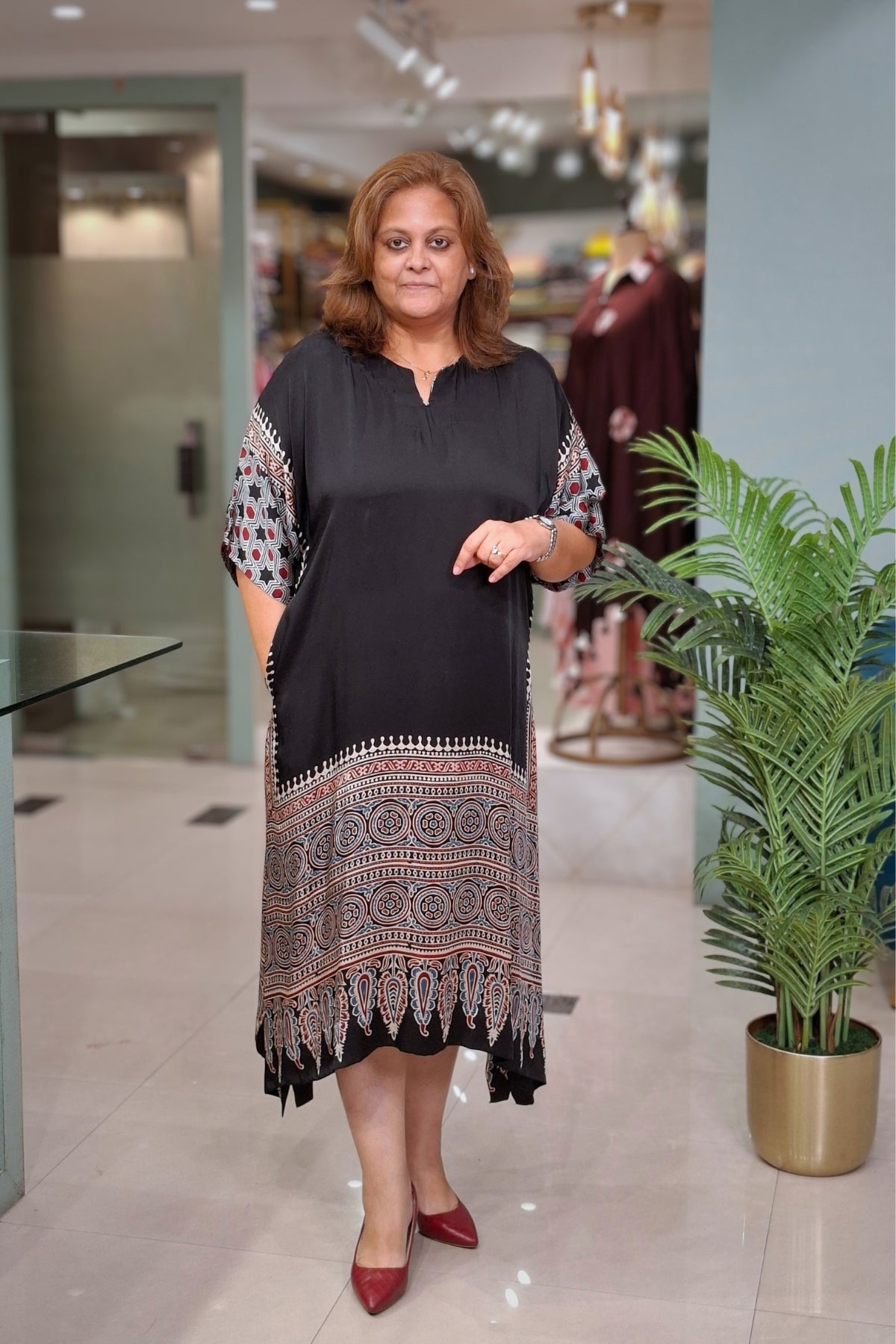 A woman modeling a black tunic dress with intricate multicolor geometric patterns on the sleeves and hem, standing in a modern indoor boutique setting.