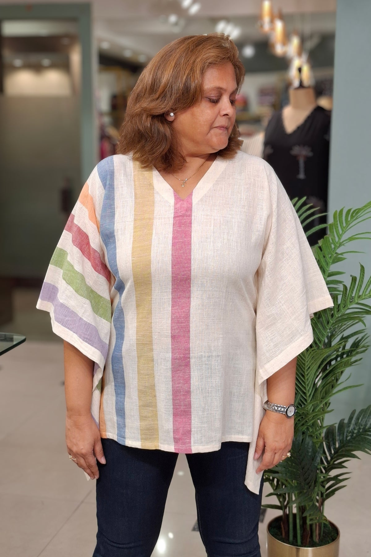 A woman wearing a loose, short-sleeved linen top with vertical pastel stripes in pink, yellow, blue, green, and purple on an off-white base, paired with dark blue jeans, standing in a store.