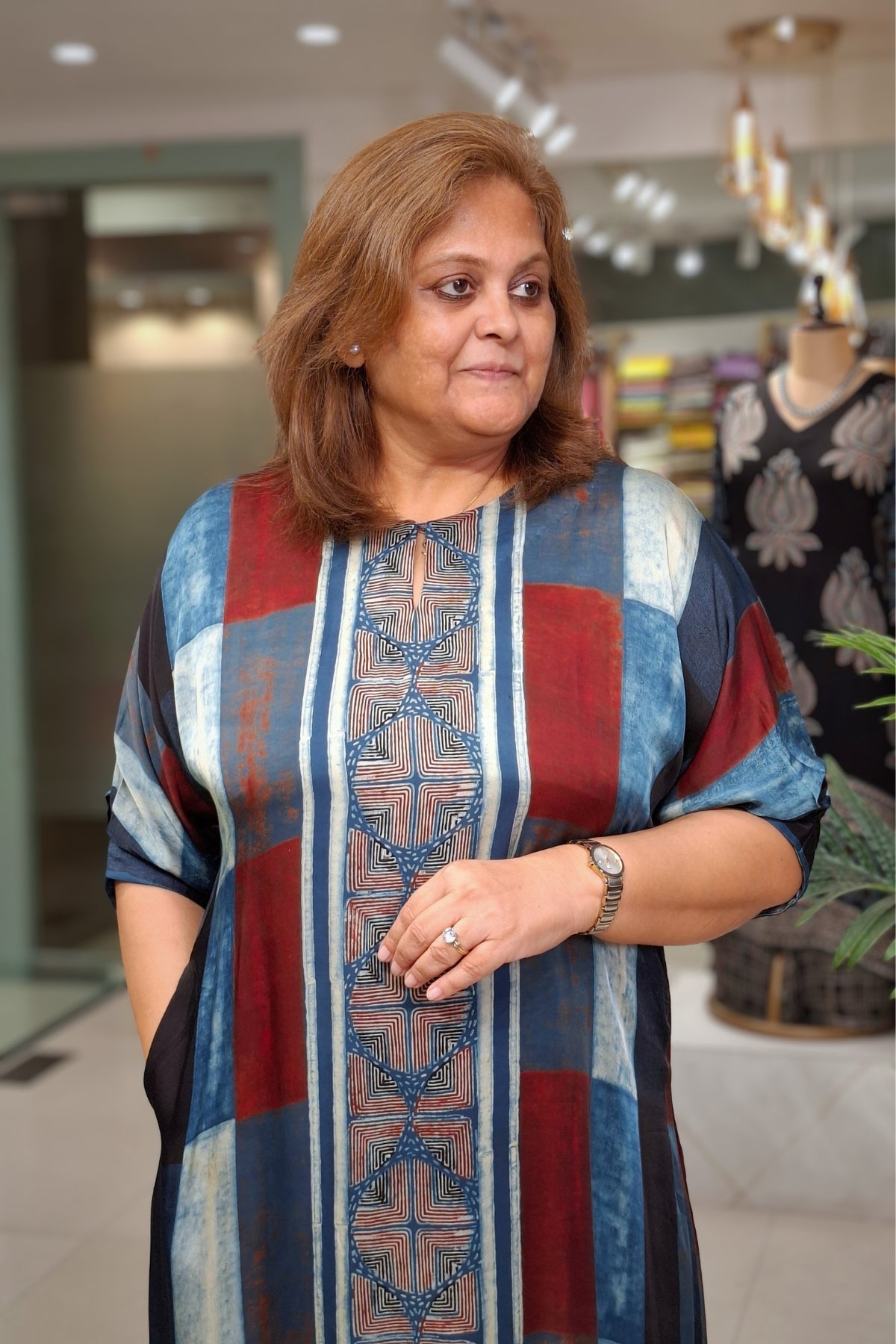 A woman wearing a loose, Elbow-sleeved tunic Dress with a patchwork design in red, blue, and beige, standing inside a clothing store.