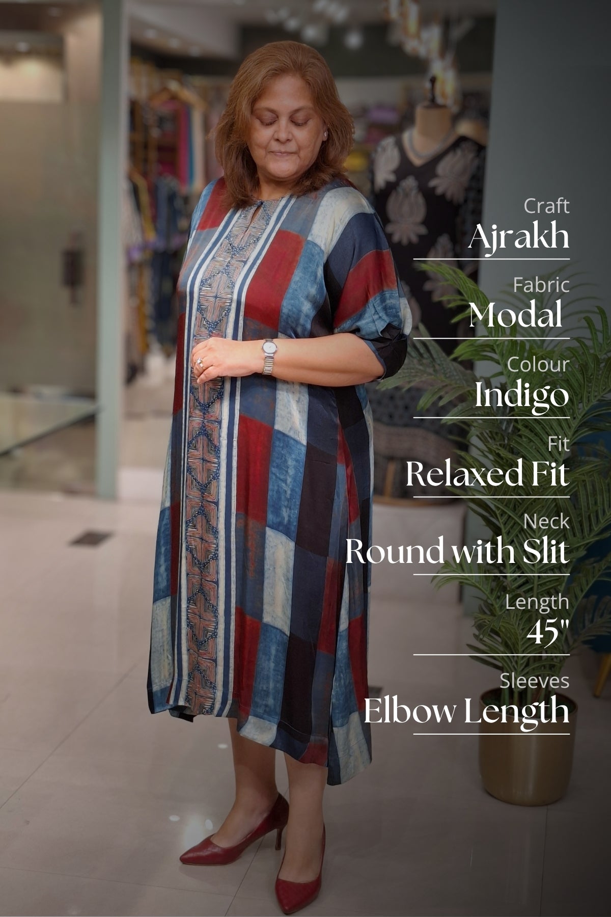 A woman modeling a long Ajrakh fabric modal dress with a patchwork pattern in blue, red, and beige, standing in a boutique. Text overlays on the right list the dress specifications.