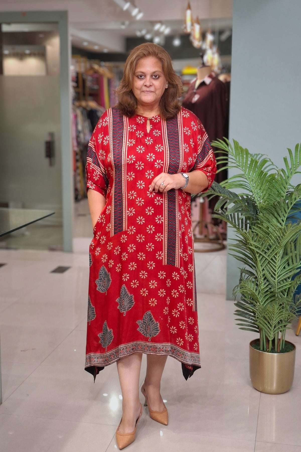 A woman modeling a red cotton kurta with white floral prints and dark green border patterns, standing in a clothing store.