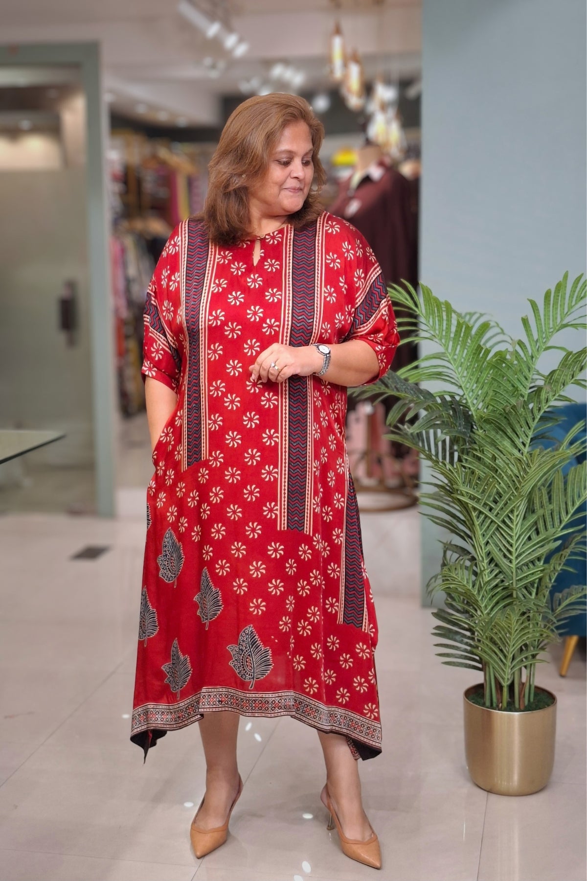 A woman modeling a red three-quarter sleeve kurta with white floral prints and dark brown vertical border panels, standing in a modern indoor setting with a potted plant in the background.