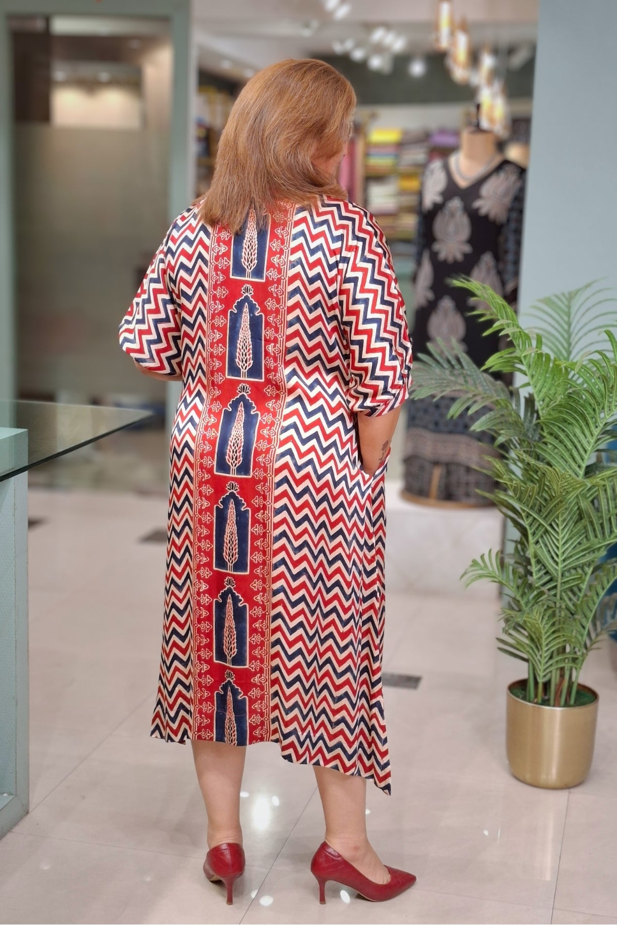 A woman viewed from behind wearing a red, navy, and white patterned dress in a boutique setting, paired with red heels.