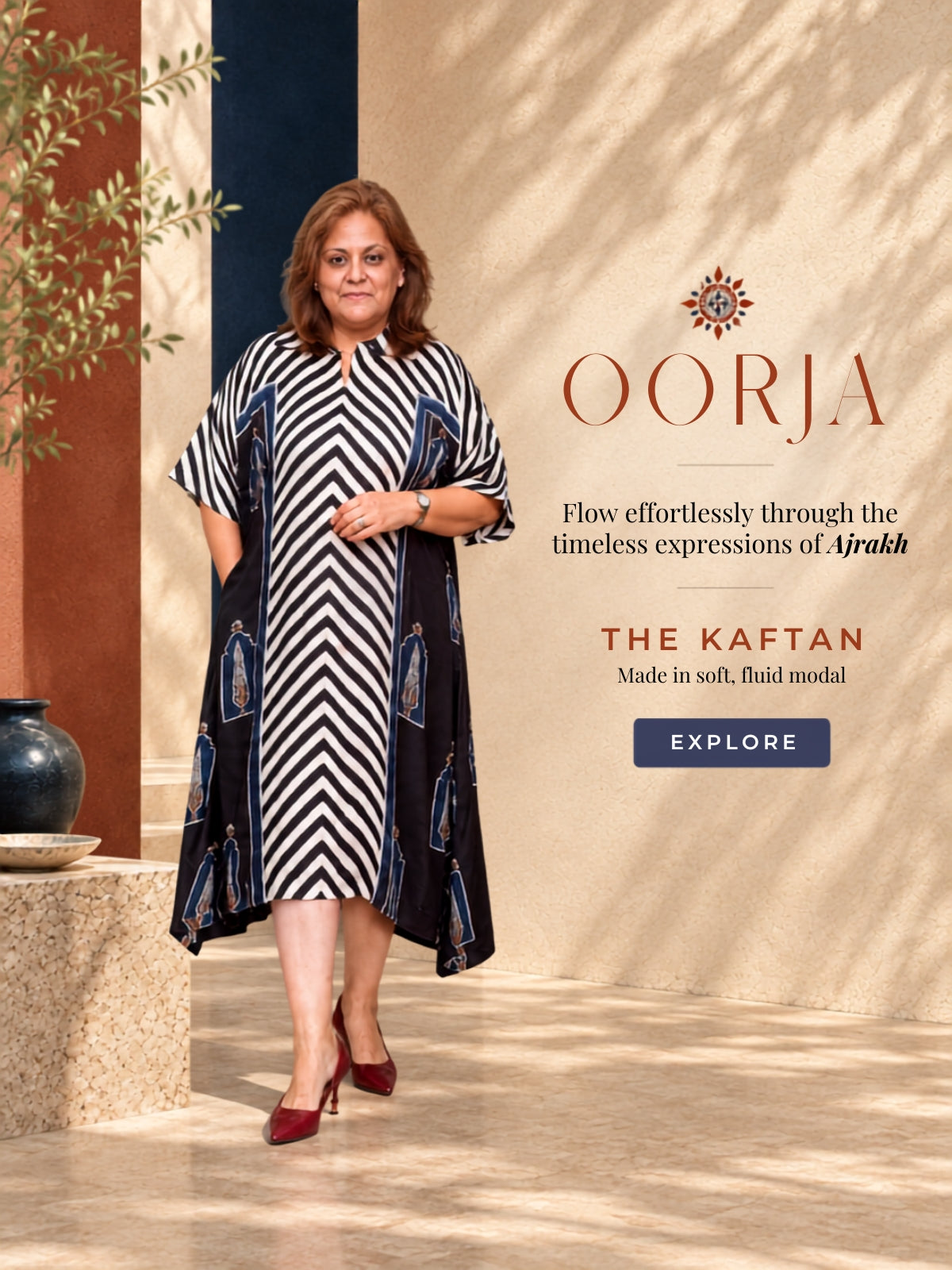 A woman modeling a black and white chevron-patterned kaftan dress with blue and gold printed border details, standing against a beige textured wall with promotional text for OORJA.