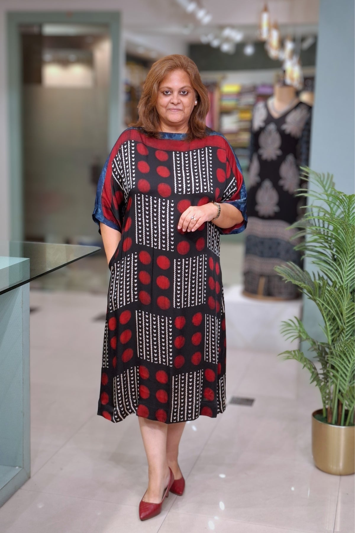 A woman wearing a loose-fitting A-line dress with a red, black, and white geometric pattern, standing in a modern indoor boutique setting.