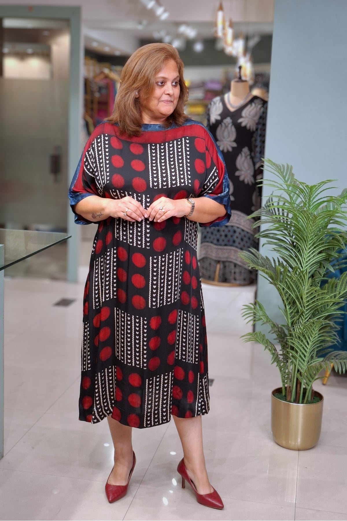 A woman wearing a tiered midi dress with a black, red, and white geometric pattern, standing in a boutique next to a potted plant.