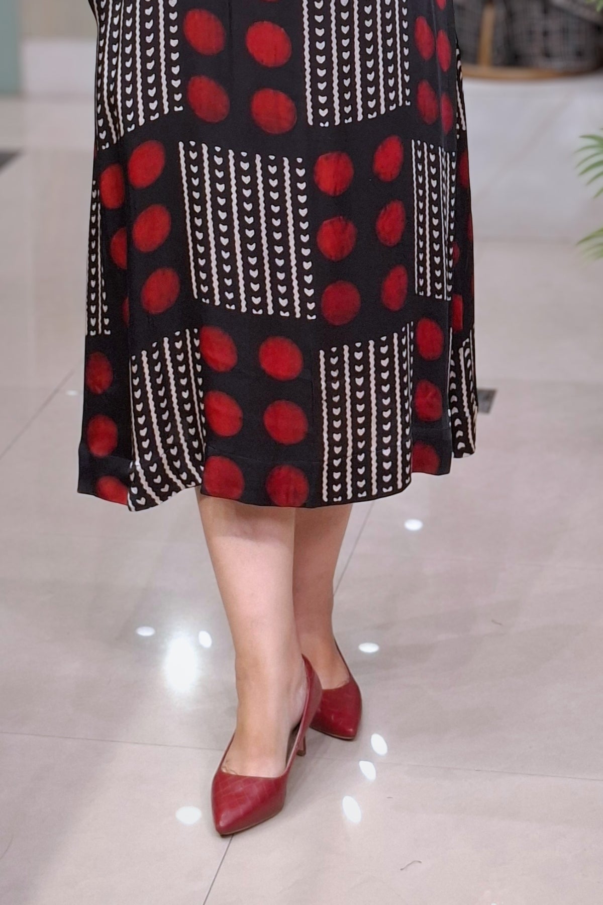 A person wearing a black midi skirt with red polka dots and vertical white patterned stripes, paired with red pointed-toe heels, standing on a light tiled floor.