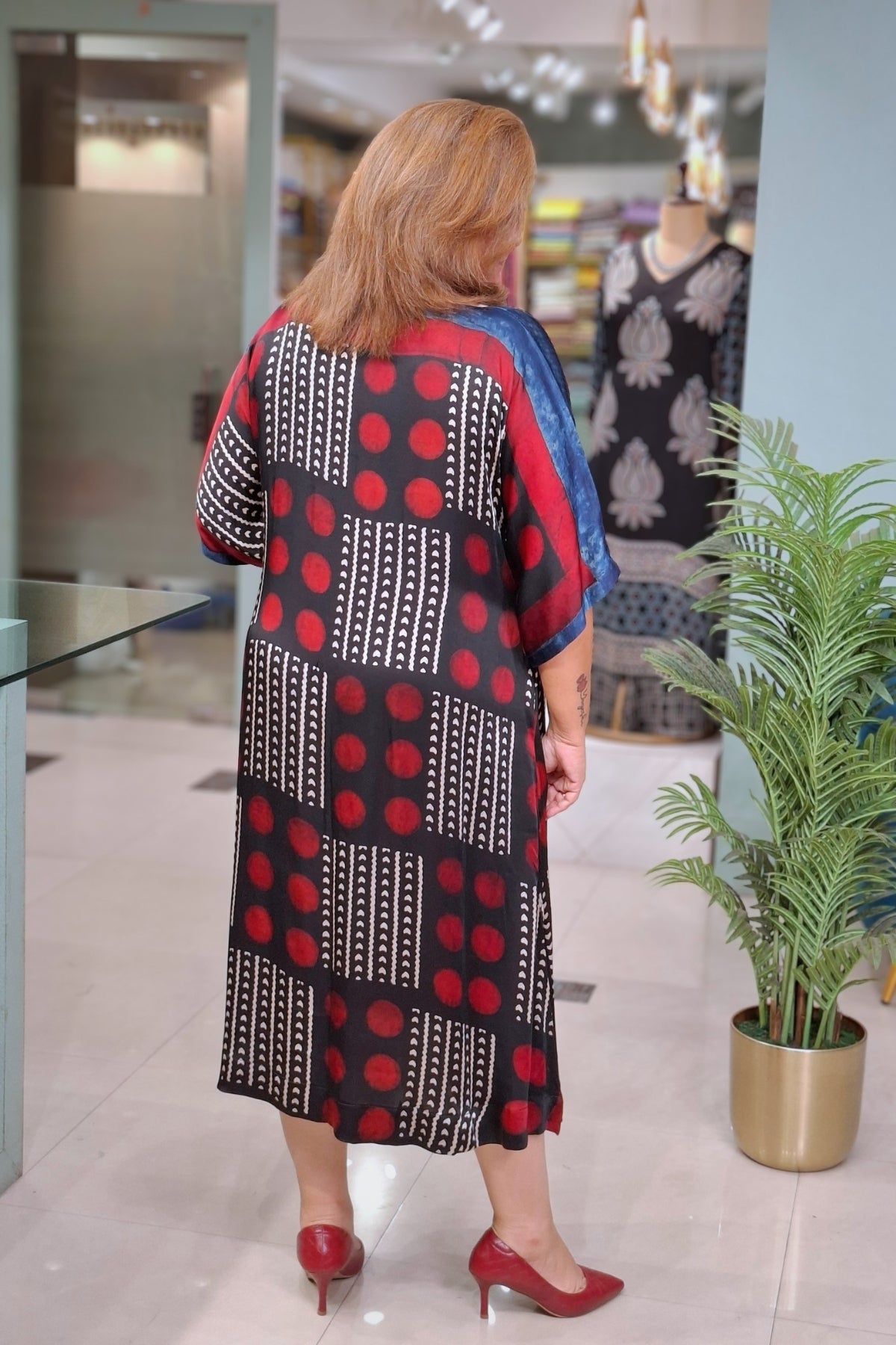 A woman viewed from behind wearing a loose, short-sleeved midi dress with a bold geometric patchwork pattern in black, red, and white, paired with red heels in a retail store setting.