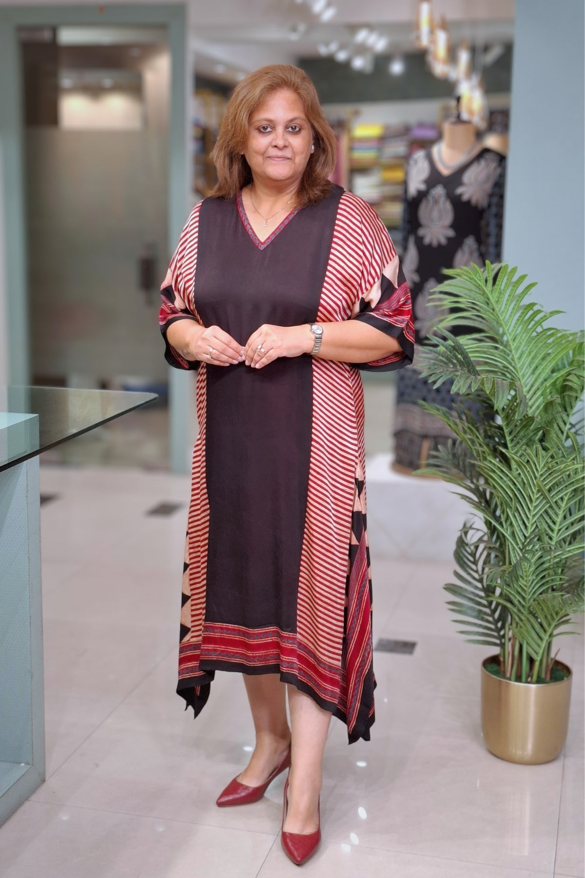 A woman wearing a long-sleeved, loose-fitting tunic dress with a dark brown body and red-and-white striped side panels, standing in a modern indoor boutique setting.