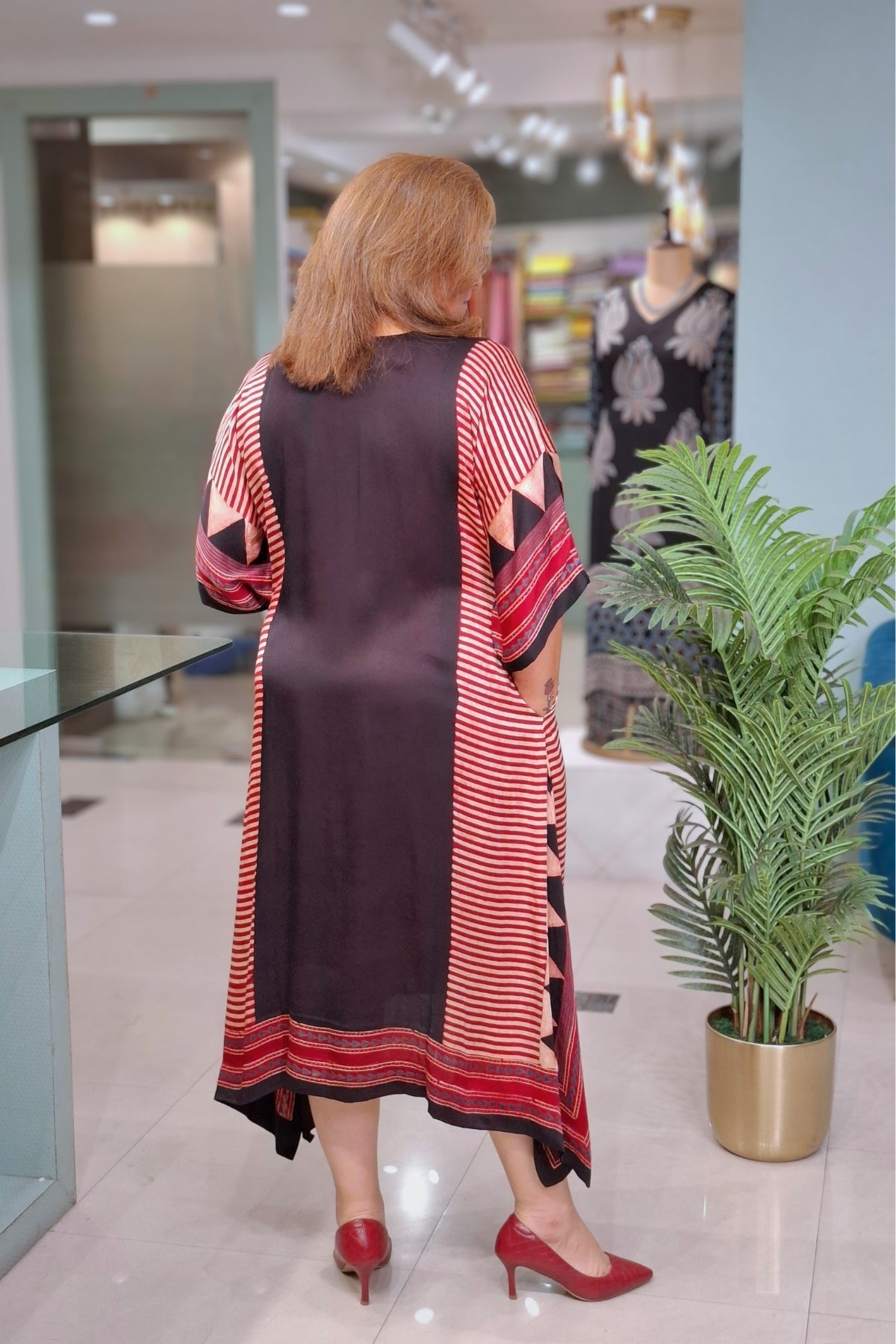 A woman viewed from behind wearing a long, loose-fitting tunic dress with a dark brown center panel and red-and-white striped sleeves and hem, standing in a modern indoor boutique next to a potted plant.