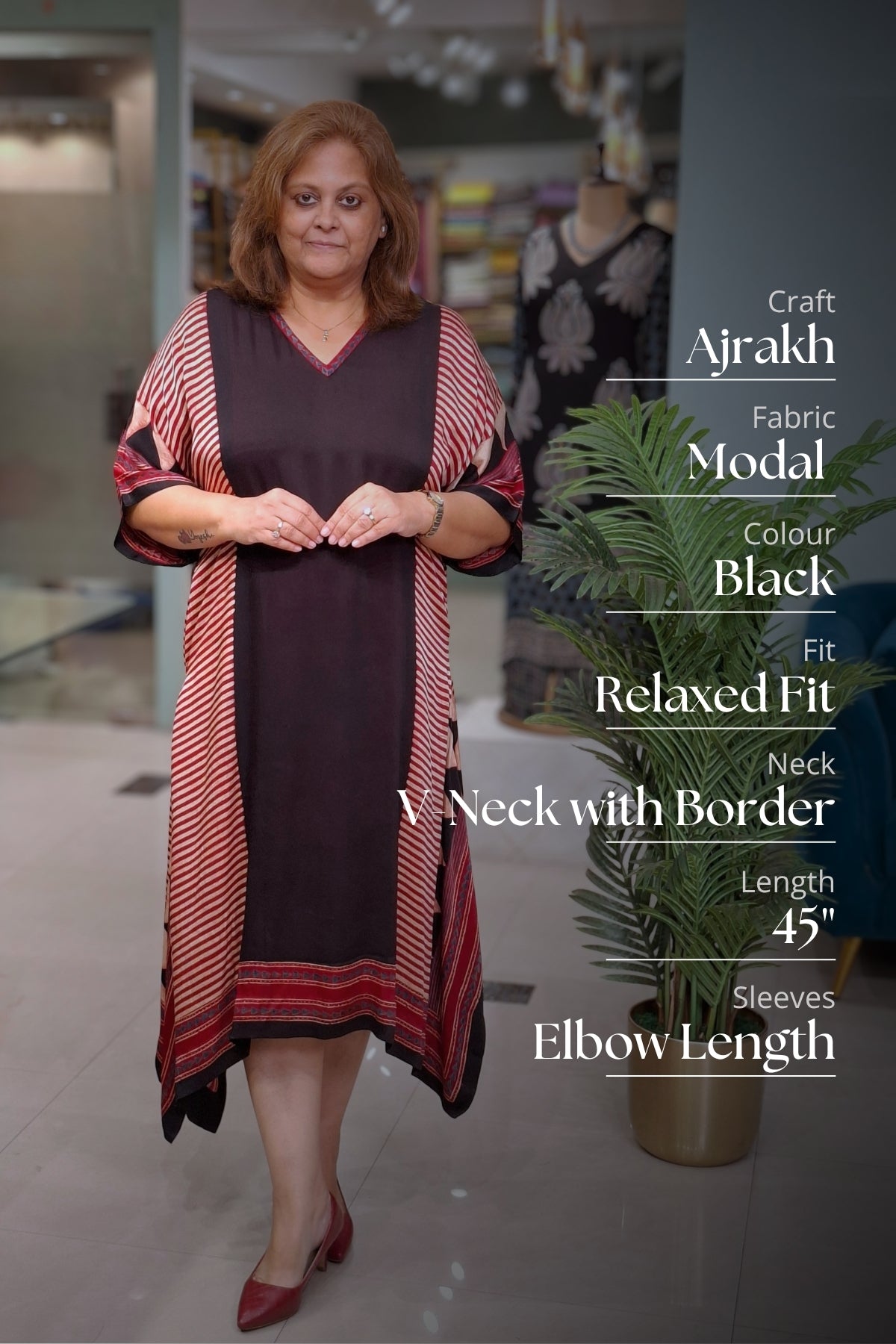 A woman modeling a black Craft Ajrakh fabric garment with red and beige striped sleeves and hem, standing in a boutique setting with product details text overlaid on the right side.