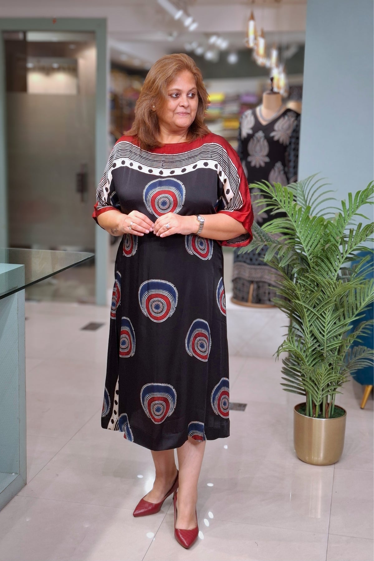 A woman modeling a black midi dress with red and blue concentric circle patterns, styled with red pointed-toe heels in a modern indoor boutique setting.