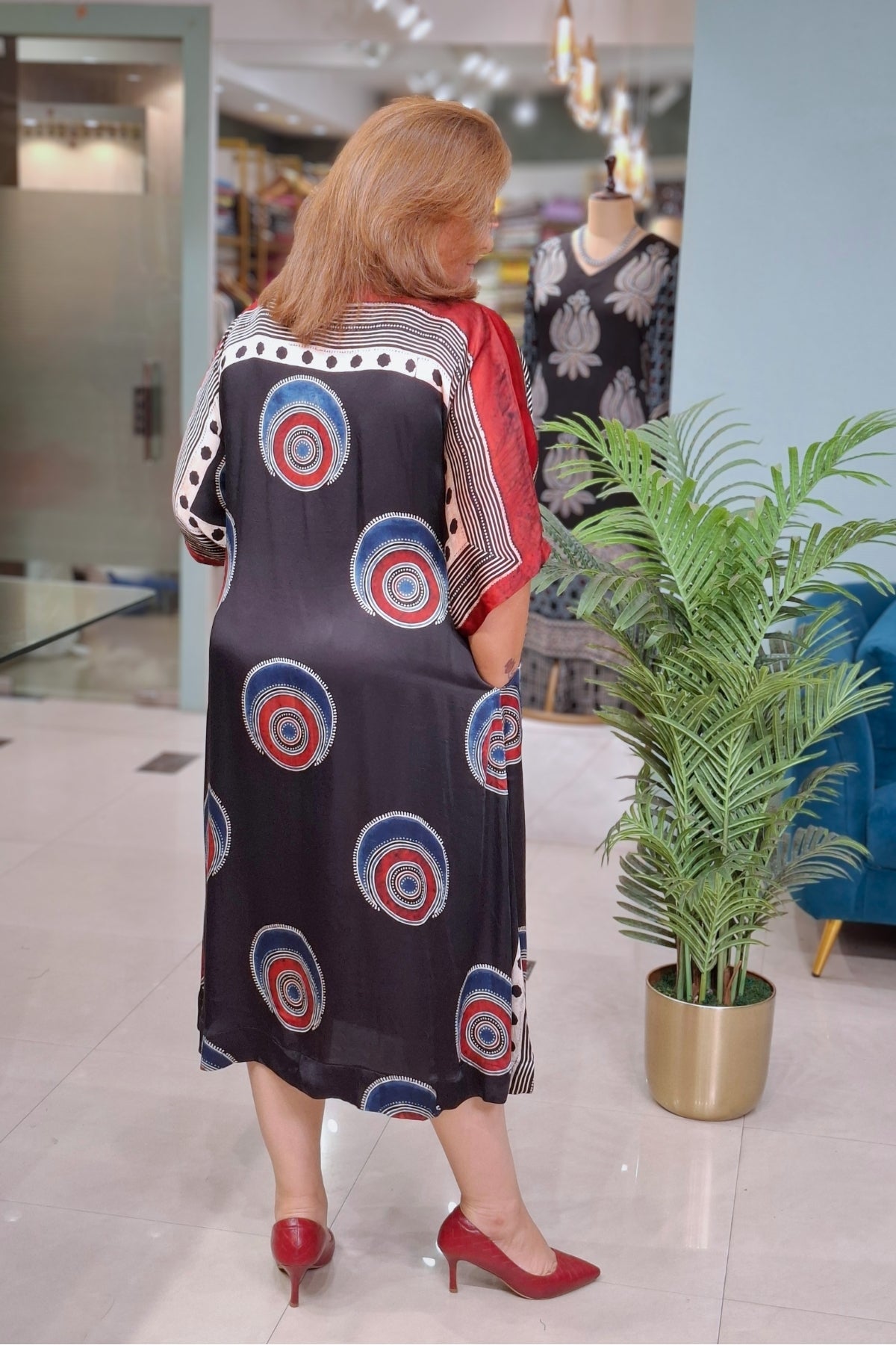 A woman viewed from behind wearing a loose, knee-length dress with a black base and circular red, white, and blue target motifs, along with red heels, standing in a boutique next to a potted plant.