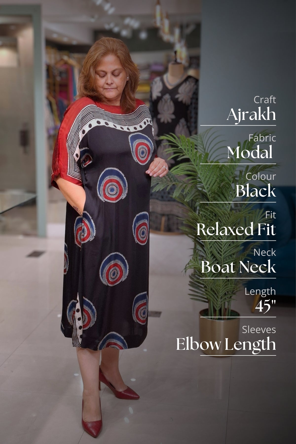 A woman modeling a black Ajrakh fabric dress with red, blue, and white circular patterns and red sleeves, standing in a store. Text on the right lists product details: Craft Ajrakh, Fabric Modal, Colour Black, Fit Relaxed Fit, Neck Boat Neck, Length 45 inches, Sleeves Elbow Length.