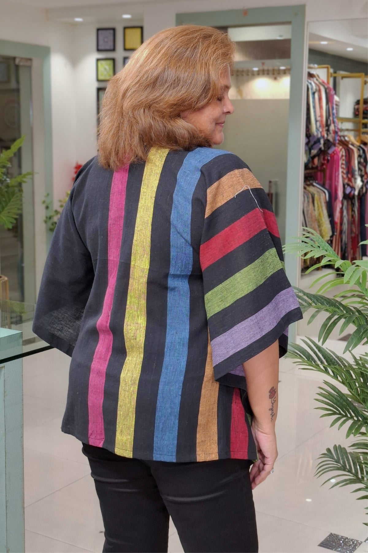 A woman wearing a black kurta with vibrant vertical stripes in yellow, blue, pink, orange, green, and purple, photographed from behind inside a clothing boutique.