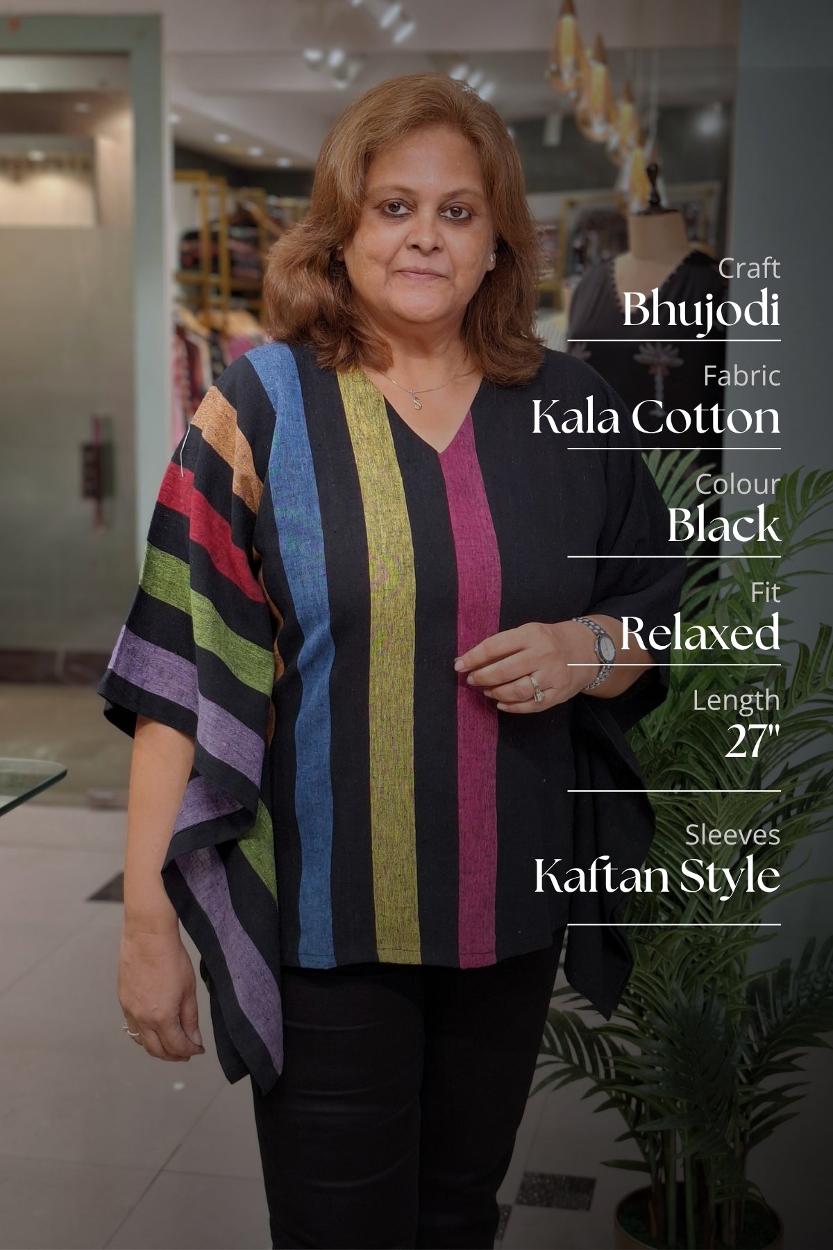 A woman modeling a black relaxed-fit Bhujodi kaftan top with vertical stripes in gold, green, red, blue, and purple, set against a blurred indoor boutique background with text overlays detailing the product specifications.