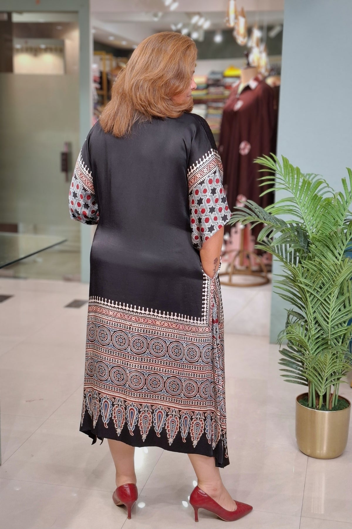 A woman viewed from behind wearing a black tunic dress with intricate multicolor geometric patterns on the sleeves and hem, standing in a clothing store next to a potted plant.