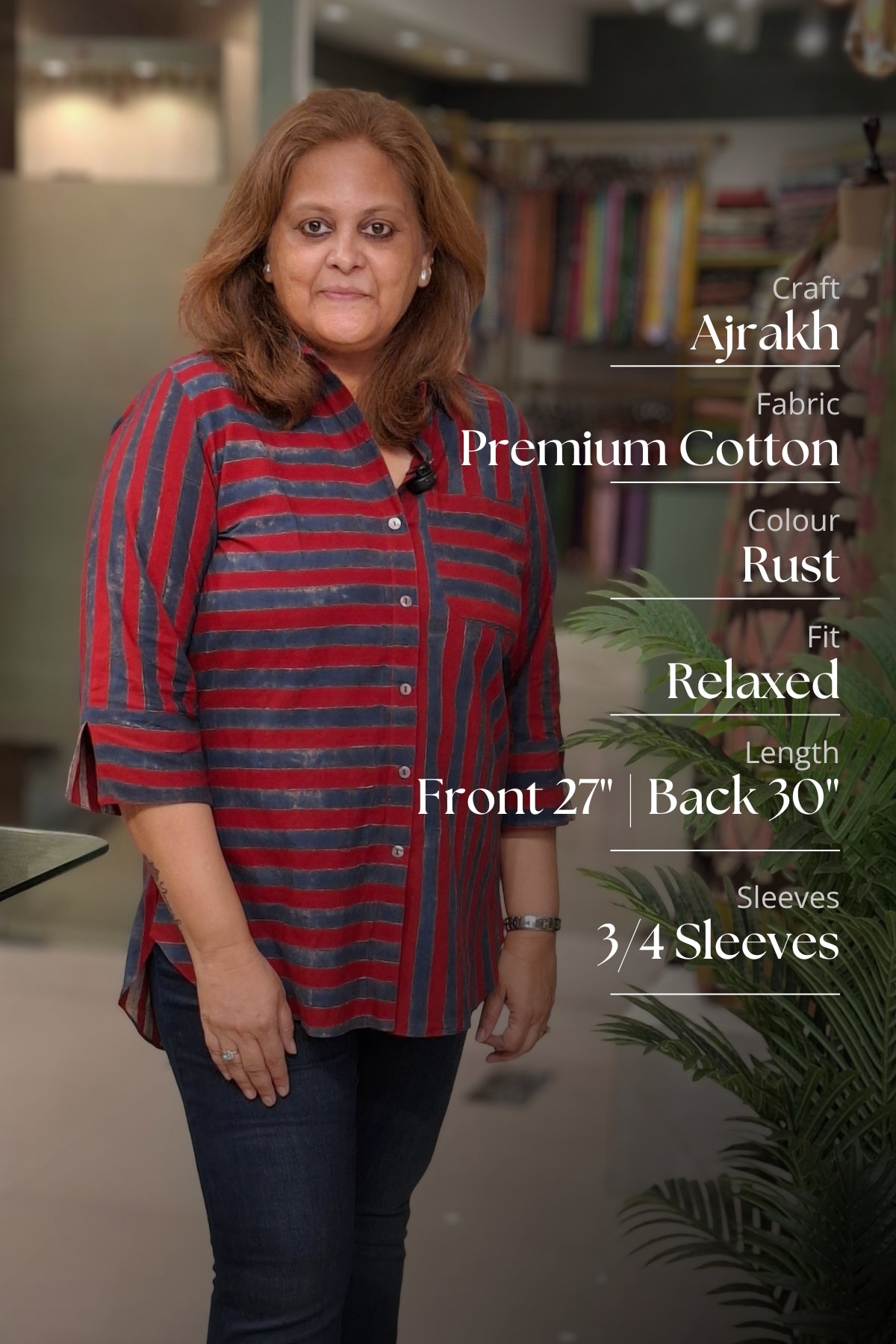 A woman modeling a red and navy striped relaxed-fit cotton shirt with dark blue jeans, standing in a store setting with text overlays detailing the product's fabric and measurements.
