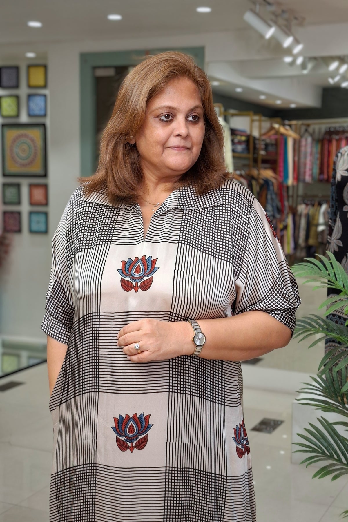 A woman wearing a black and white checkered tunic top with red and blue lotus flower motifs, standing inside a clothing store.