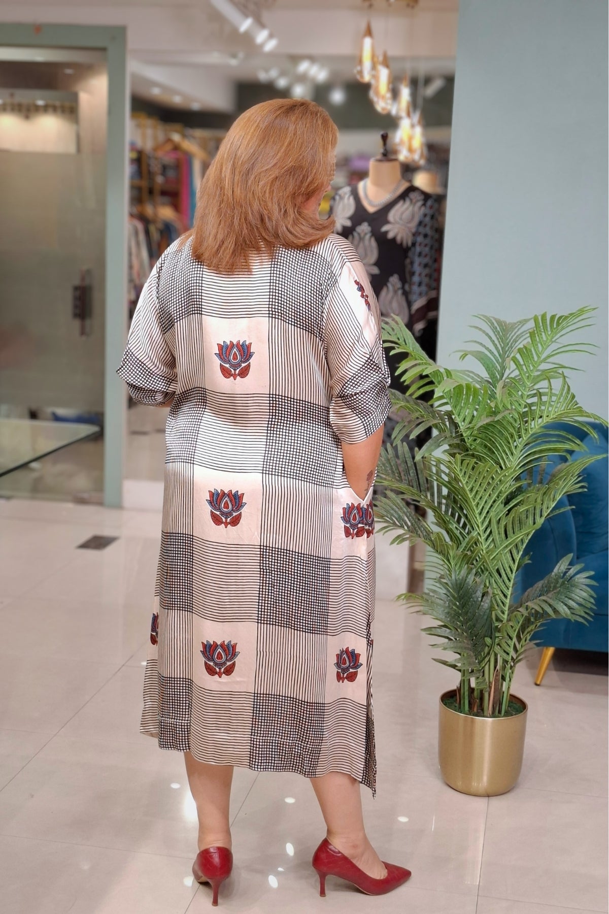 A woman viewed from behind wearing a long patchwork tunic dress with a gray, beige, and white checkered pattern and red lotus motifs, paired with red heels in a boutique setting.