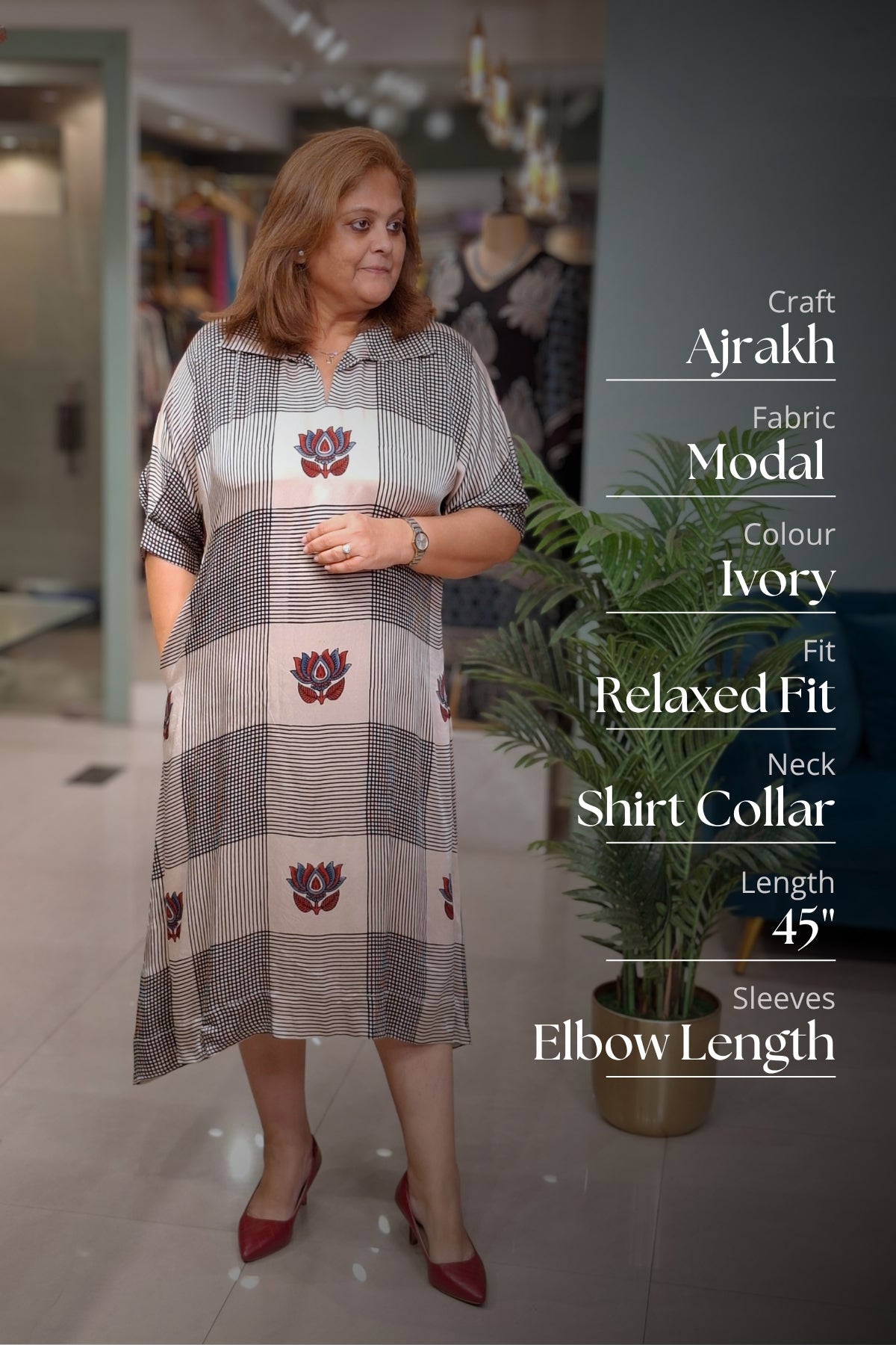 A woman modeling a gray and ivory striped Ajrakh fabric dress with red floral motifs, standing in a boutique setting with text overlays detailing the product's features.