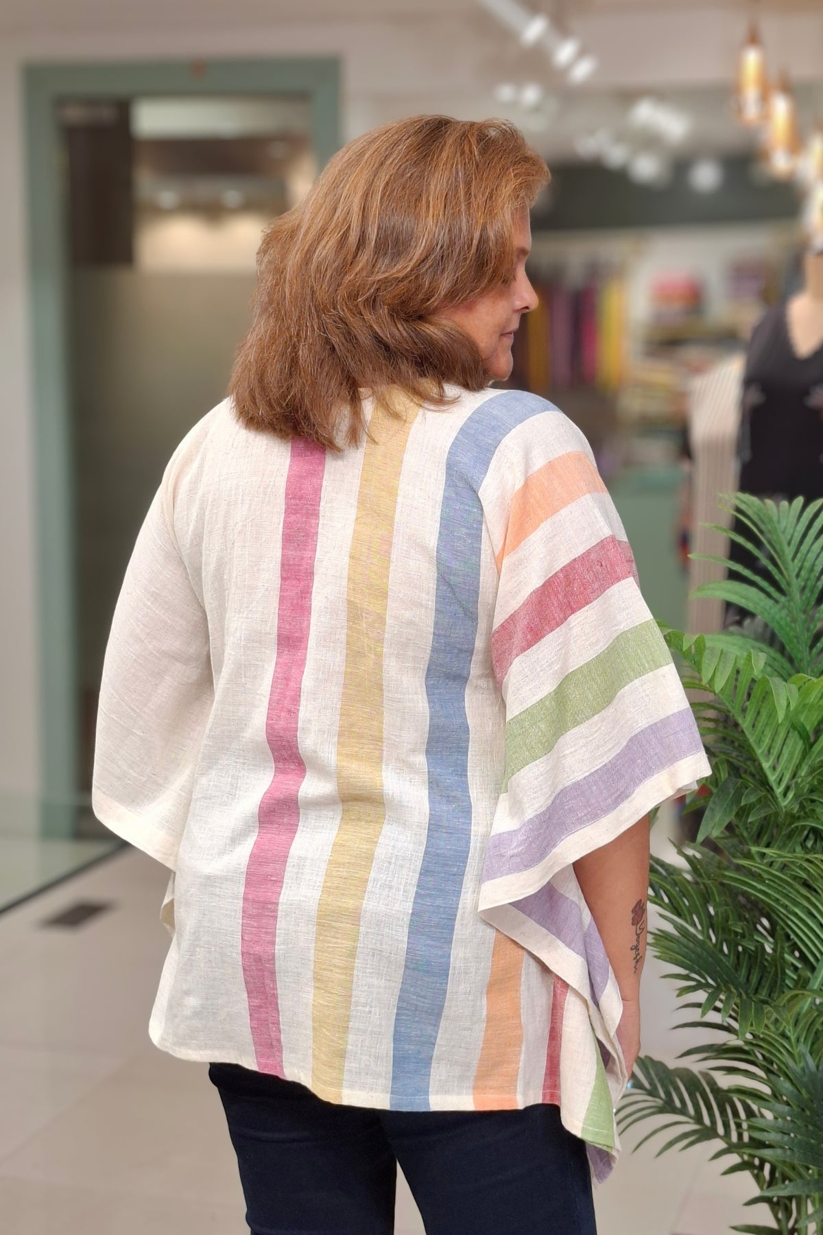 A woman viewed from behind wearing a loose white linen top with vertical pastel stripes in pink, yellow, blue, orange, and green, paired with dark pants in a store setting.