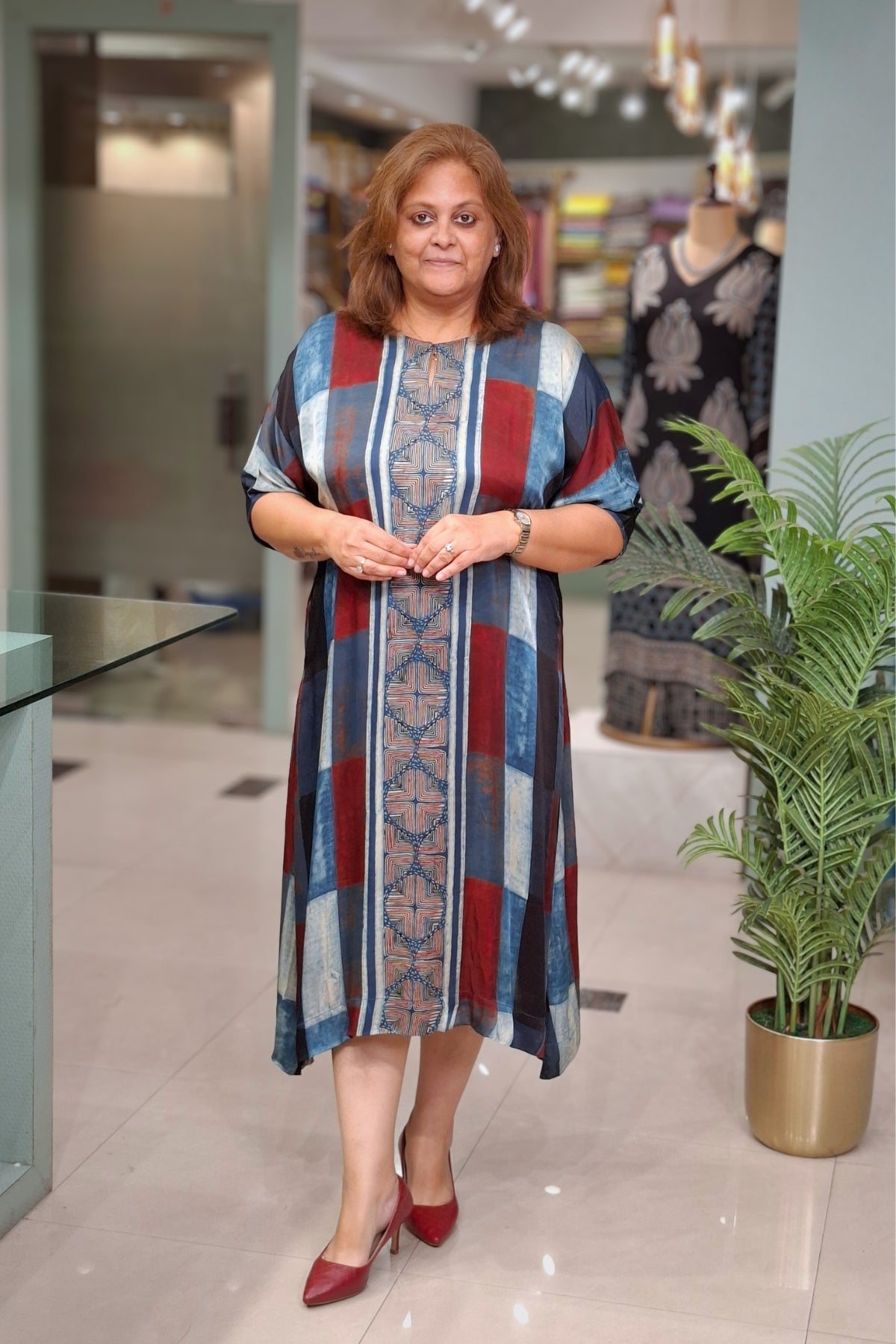 A woman wearing a loose, short-sleeved midi dress with a vertical geometric stripe pattern in blue, red, beige, and white, standing in a modern clothing store.