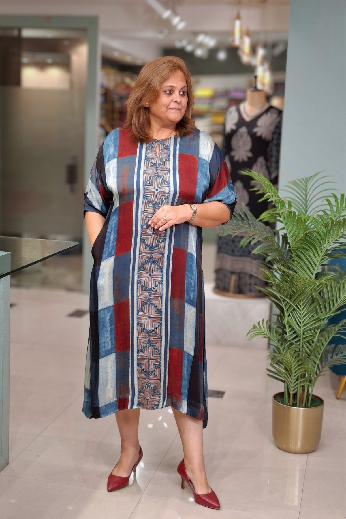 A woman modeling a loose, knee-length tunic dress with vertical stripes and geometric patterns in blue, red, beige, and white, paired with red heels, standing in a boutique setting.