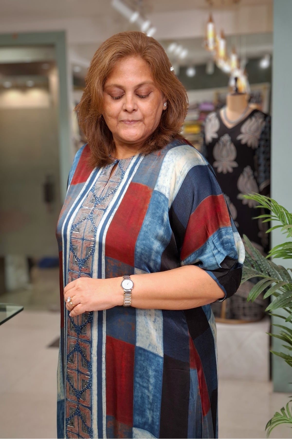 A woman wearing a colorful patchwork tunic dress with blue, red, and beige patterns, standing in a boutique.