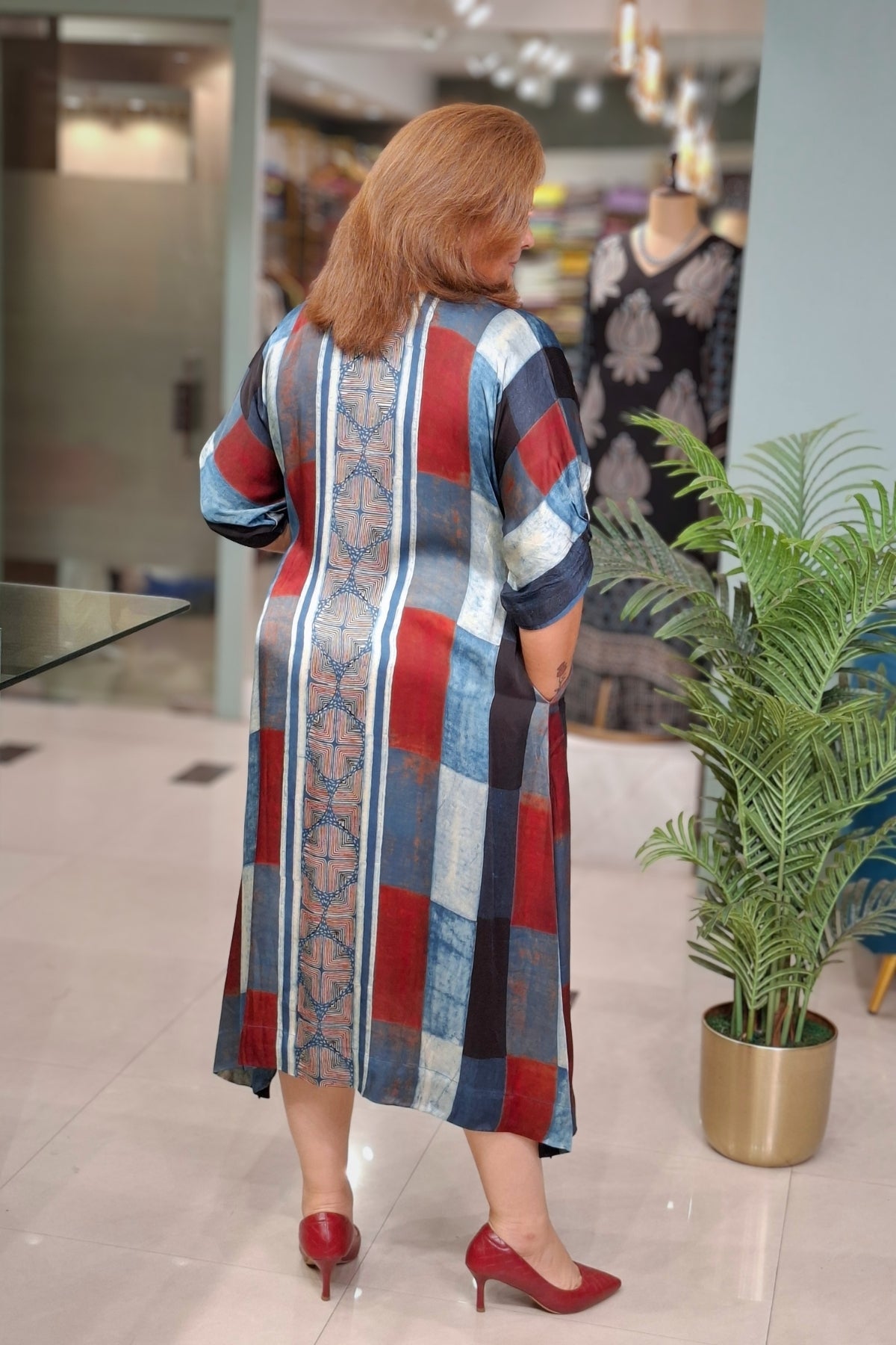 A woman viewed from behind wearing a long-sleeved patchwork midi dress with vertical stripes in red, blue, beige, and black, paired with red heels in a clothing store.