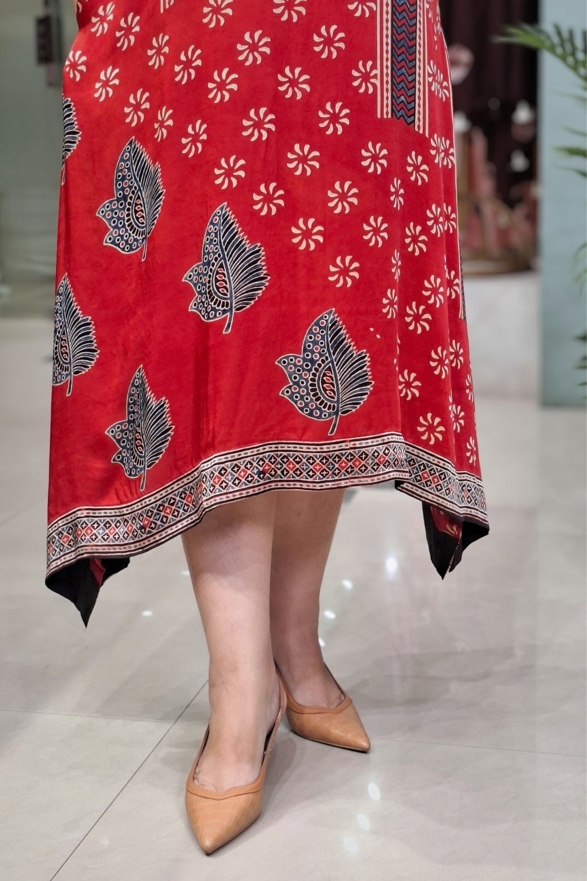 A red high-low dress with a white floral pattern and black leaf motifs, shown on a model wearing beige pointed-toe heels.