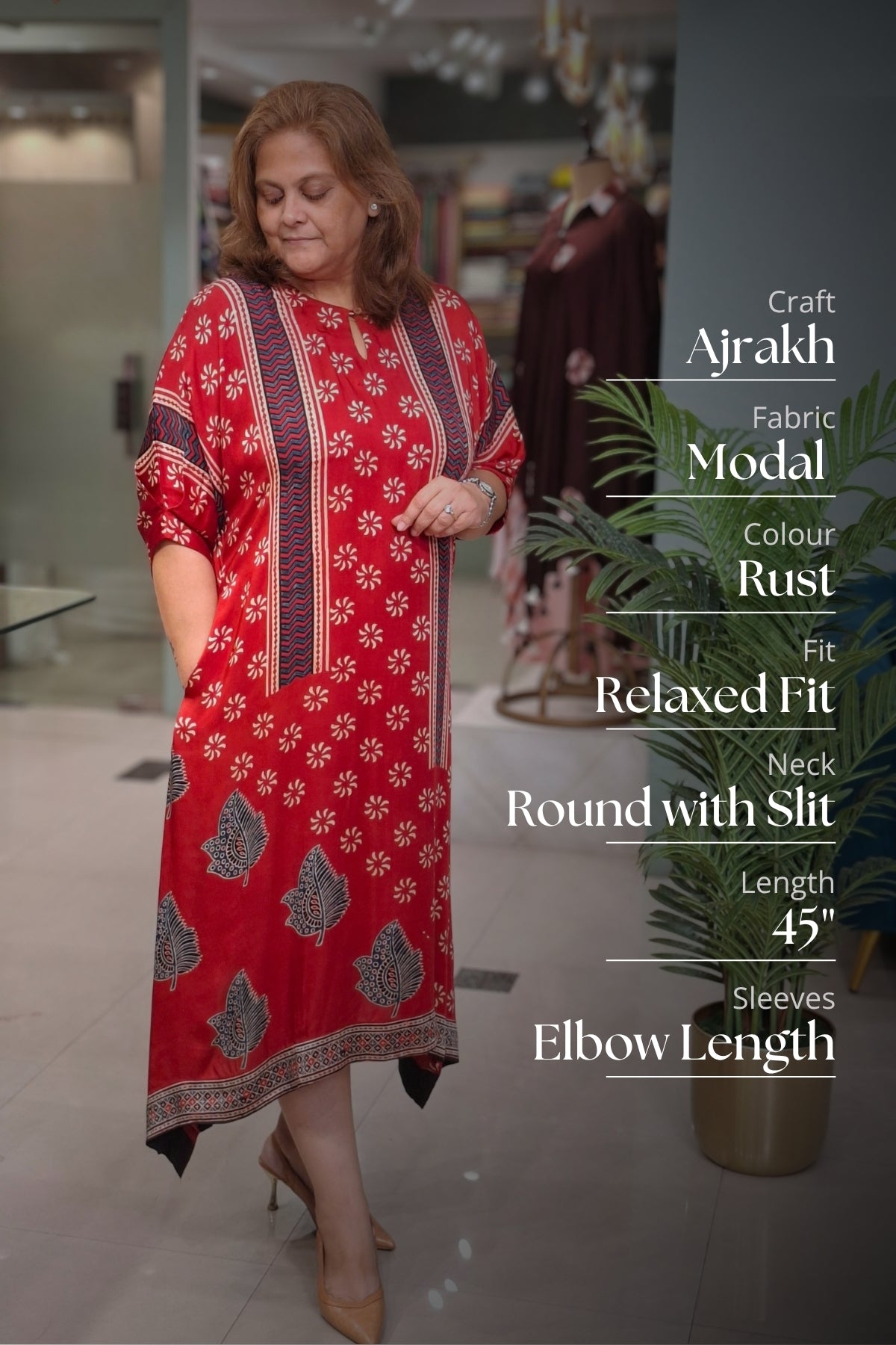 A woman modeling a rust-red Ajrakh fabric modal kurta with white floral motifs and dark patterned borders, standing in a boutique setting with text overlays detailing the garment's features.