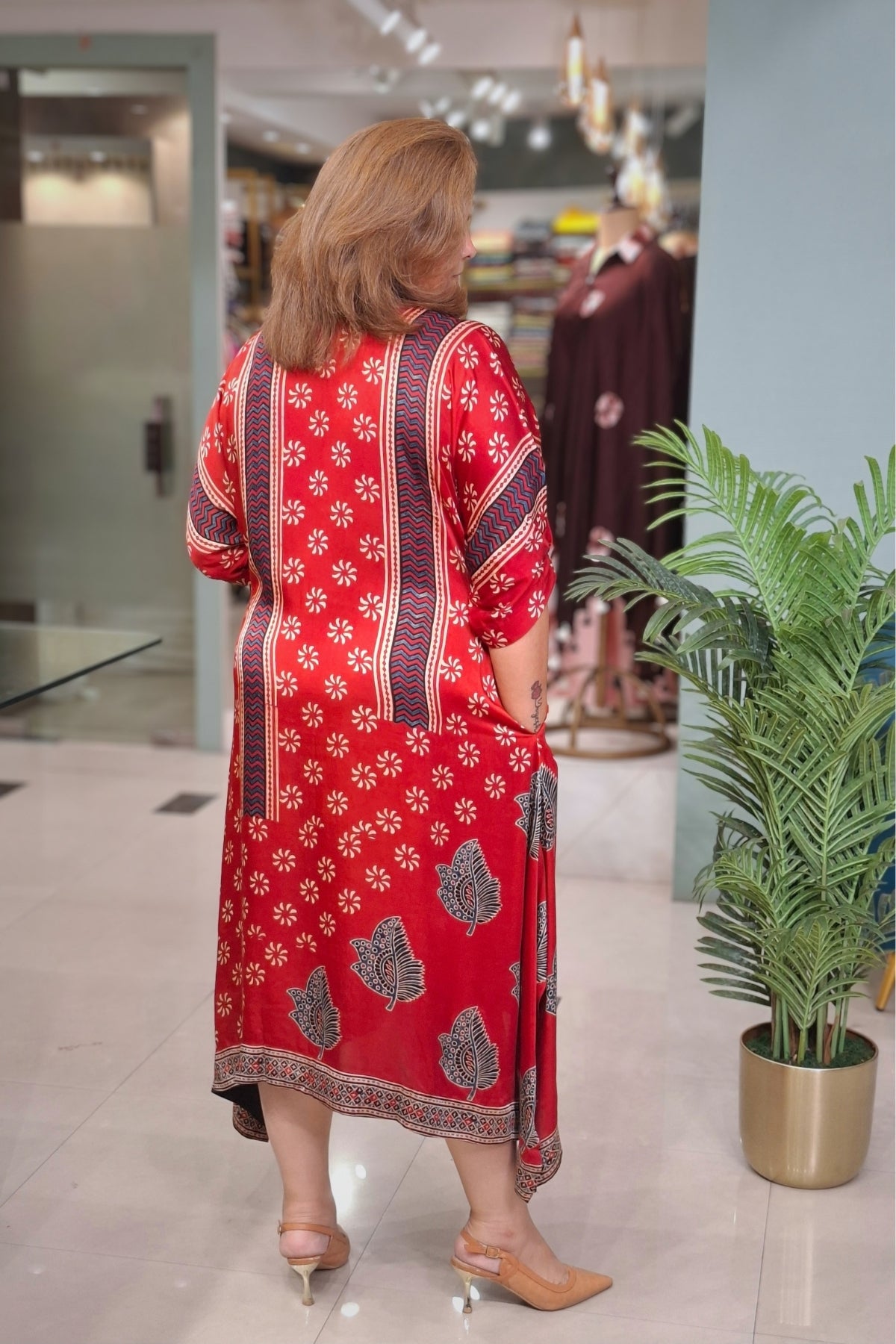 A woman viewed from behind wearing a red three-quarter sleeve kurta with white floral motifs and dark patterned borders, standing in a clothing store next to a potted plant.