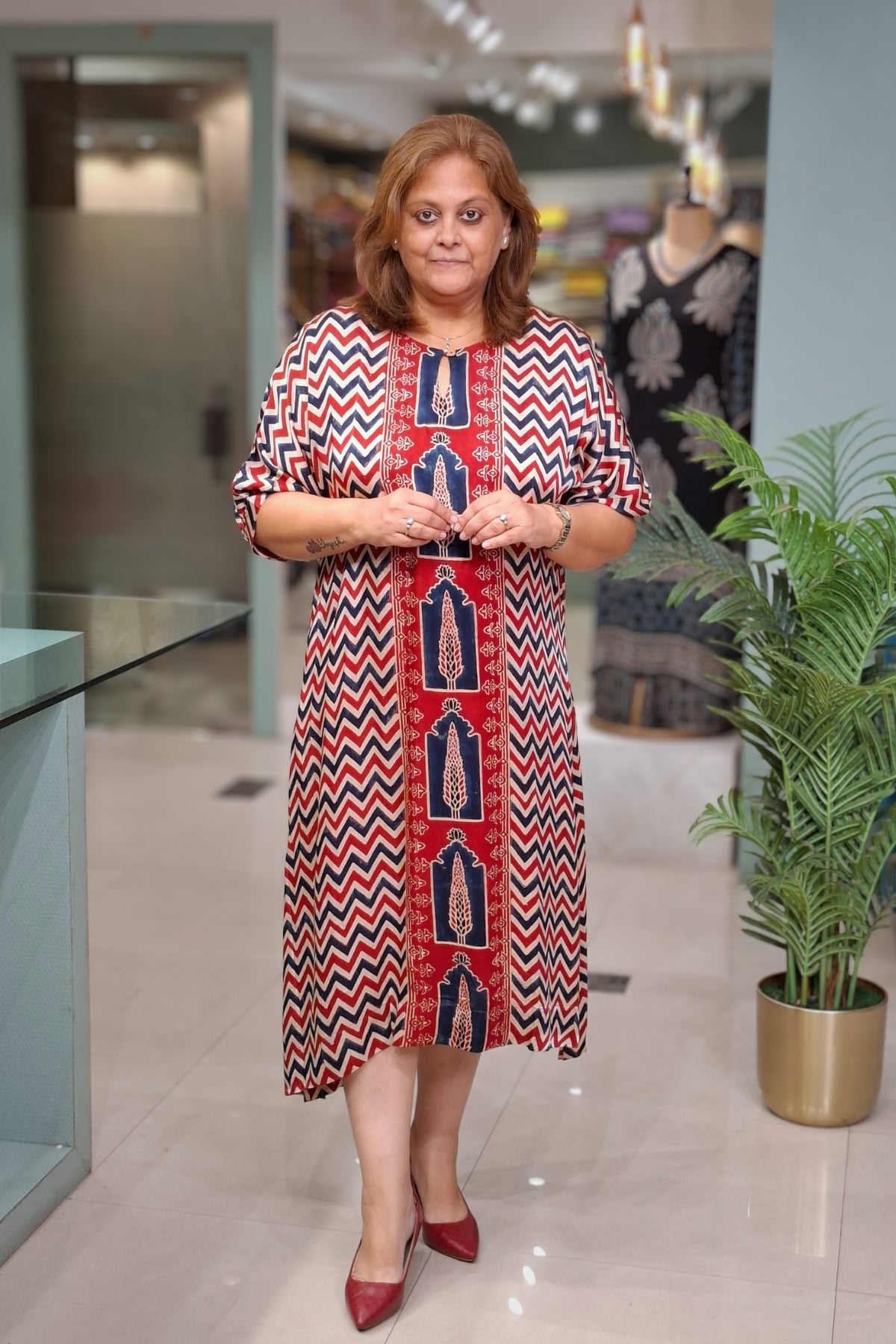 A woman wearing a red, navy, and beige chevron-patterned dress with vertical embroidered panels, standing in a retail store.