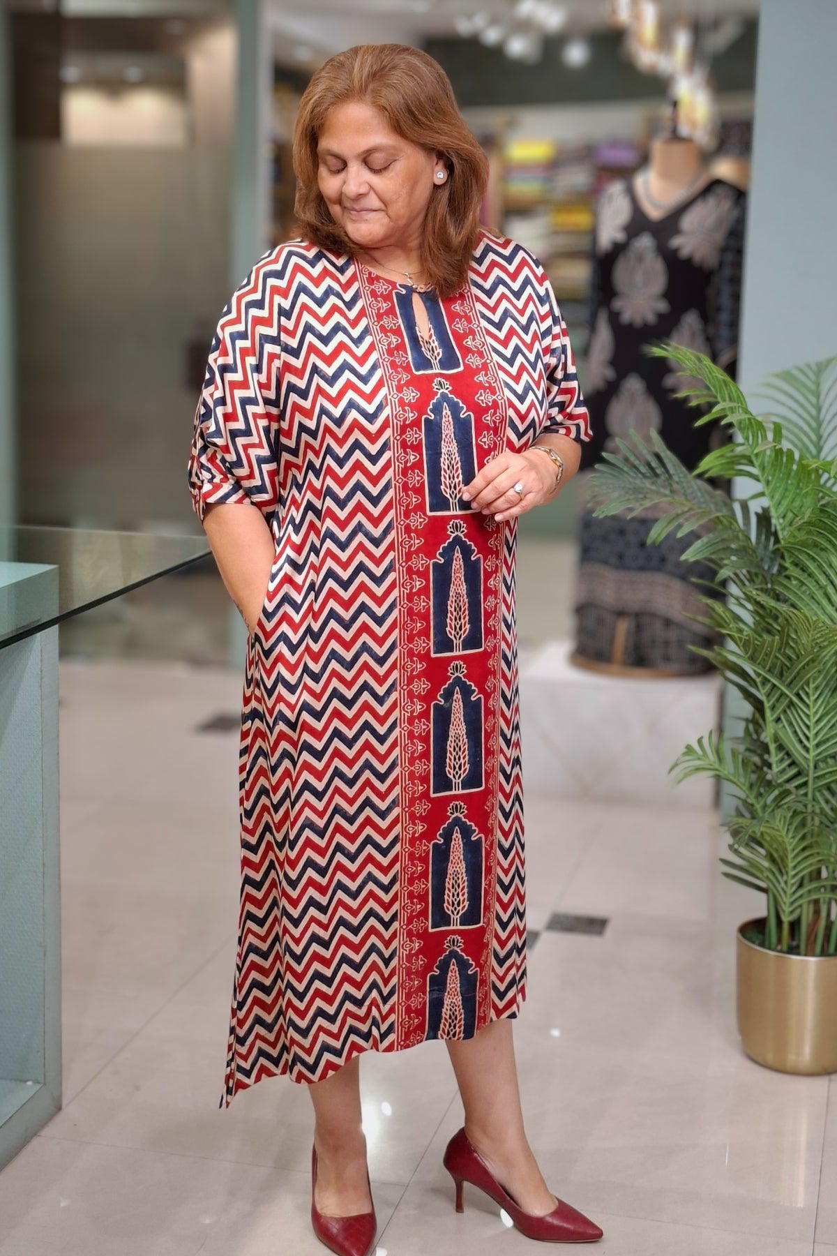 A woman wearing a red, navy, and beige chevron-patterned tunic dress with a vertical embroidered panel, standing in a modern clothing store.