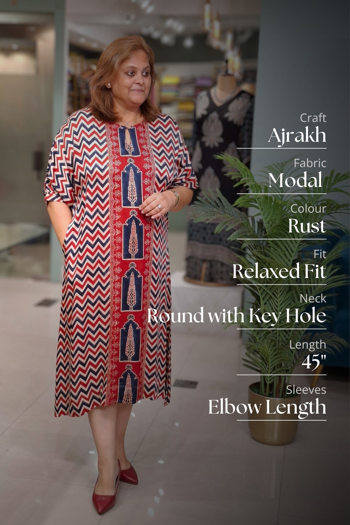 A woman modeling a rust-colored Ajrakh fabric dress with a red, navy, and beige chevron pattern and central vertical embroidery. The image features text overlays detailing the dress specifications on the right side, set in a boutique environment with a potted plant in the background.