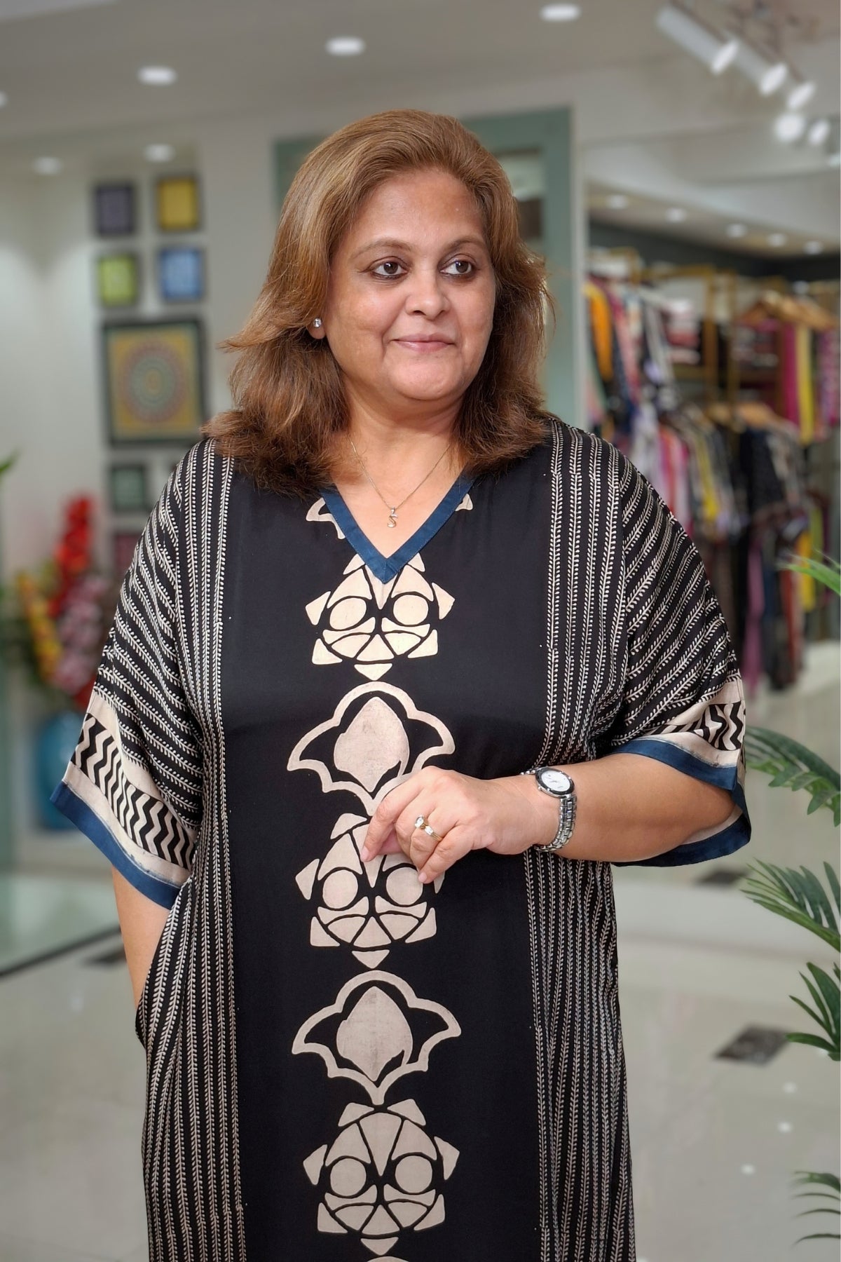 A woman wearing a black and beige patterned kaftan dress with blue trim, standing in a clothing boutique.
