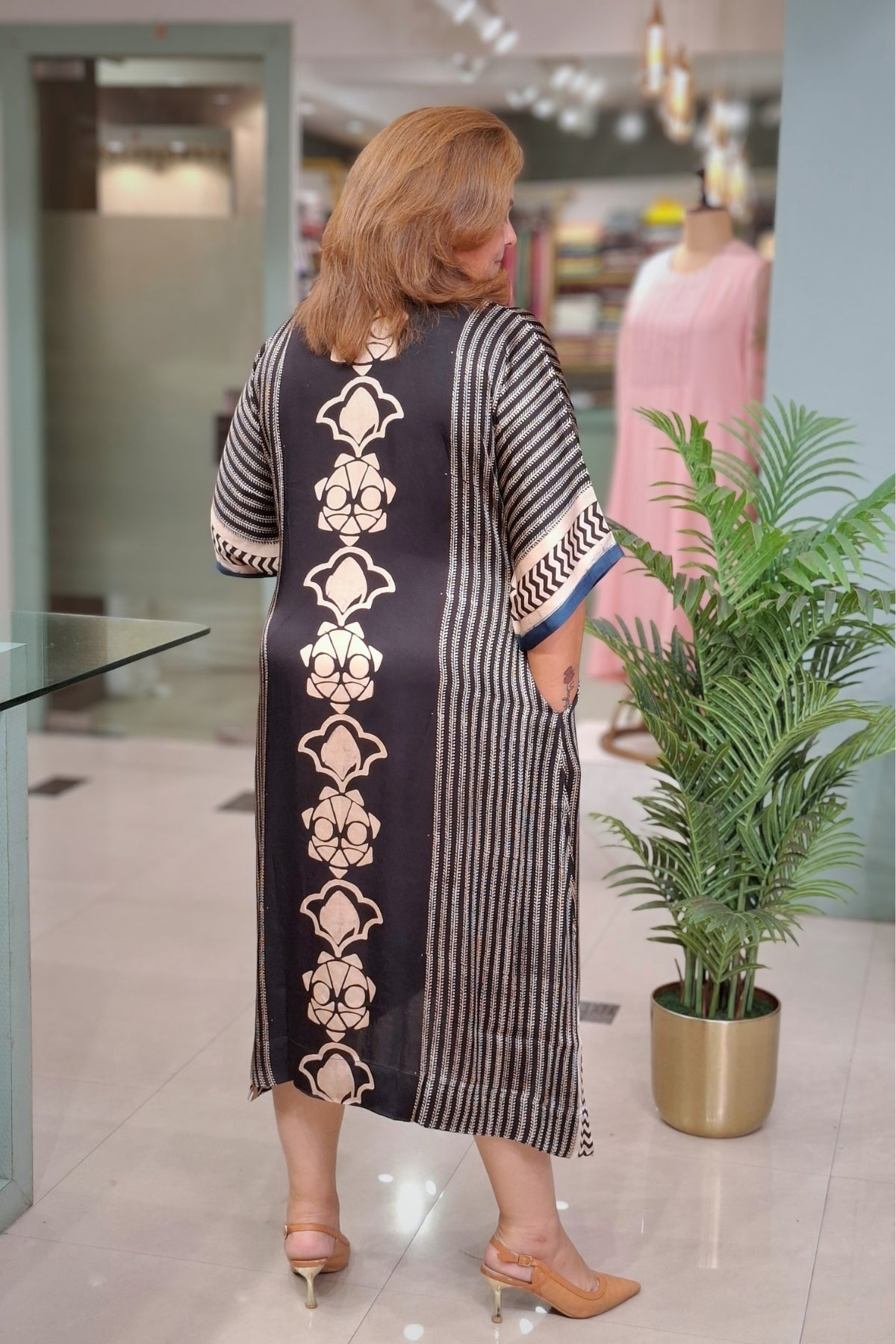 A woman wearing a black and white striped kaftan dress with beige floral patterns, photographed from behind in a boutique setting.