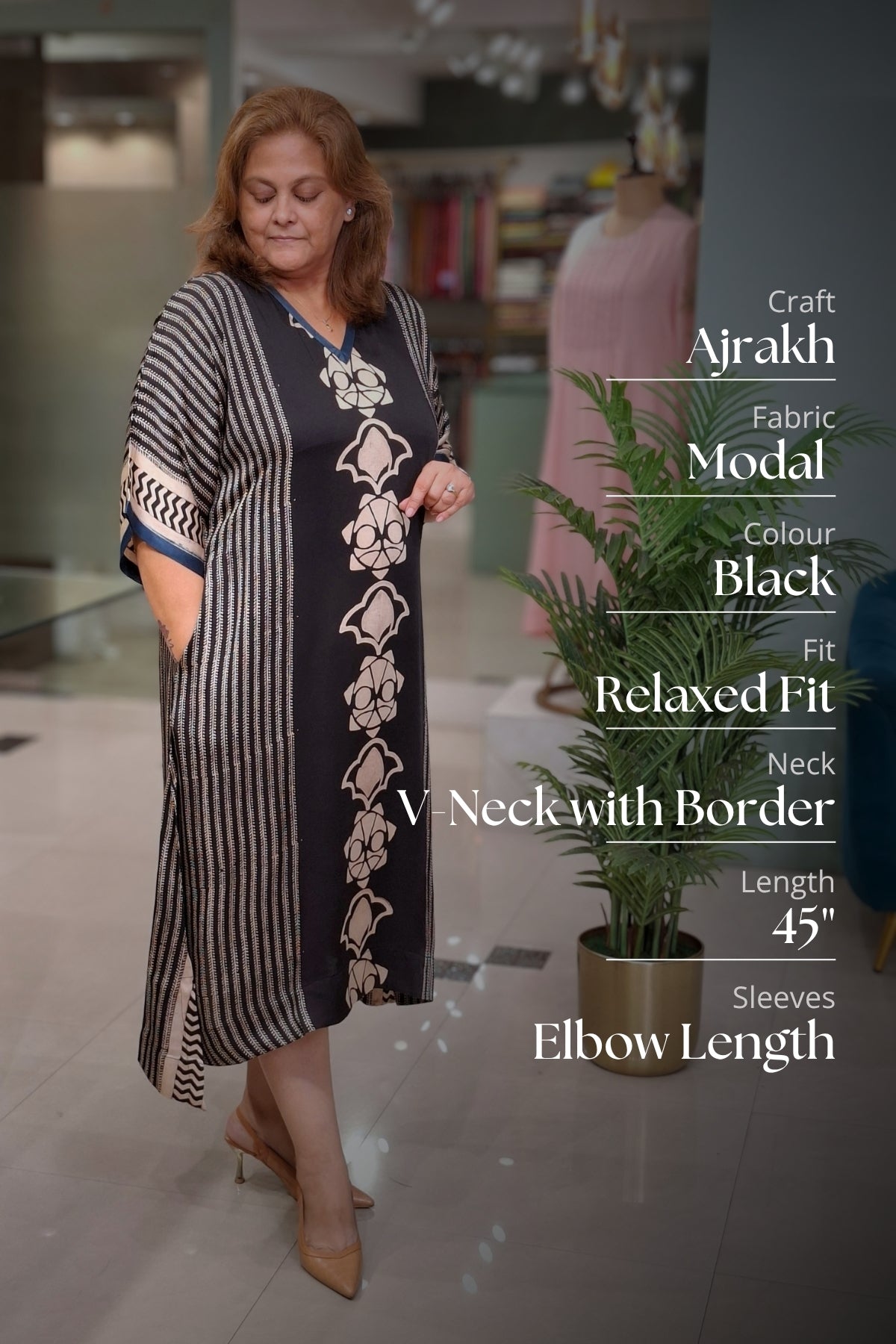 A woman modeling a black and white striped Ajrakh modal fabric dress with cream floral embroidery down the front, standing in a boutique setting with text overlays detailing the product specifications.