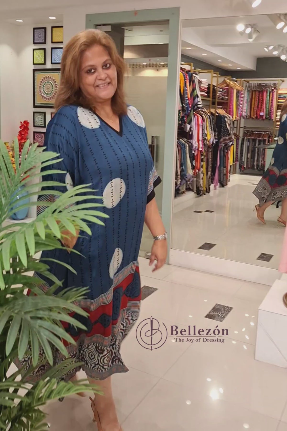 VIdeo of A woman modeling a blue kaftan dress with white circular motifs, vertical black stripes, and a red and beige patterned border at the bottom, standing in a boutique next to a potted plant.