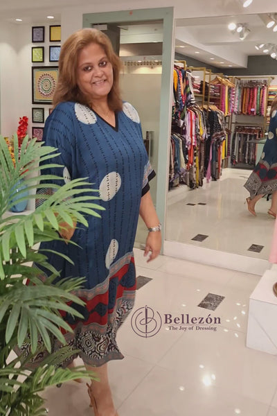 VIdeo of A woman modeling a blue kaftan dress with white circular motifs, vertical black stripes, and a red and beige patterned border at the bottom, standing in a boutique next to a potted plant.